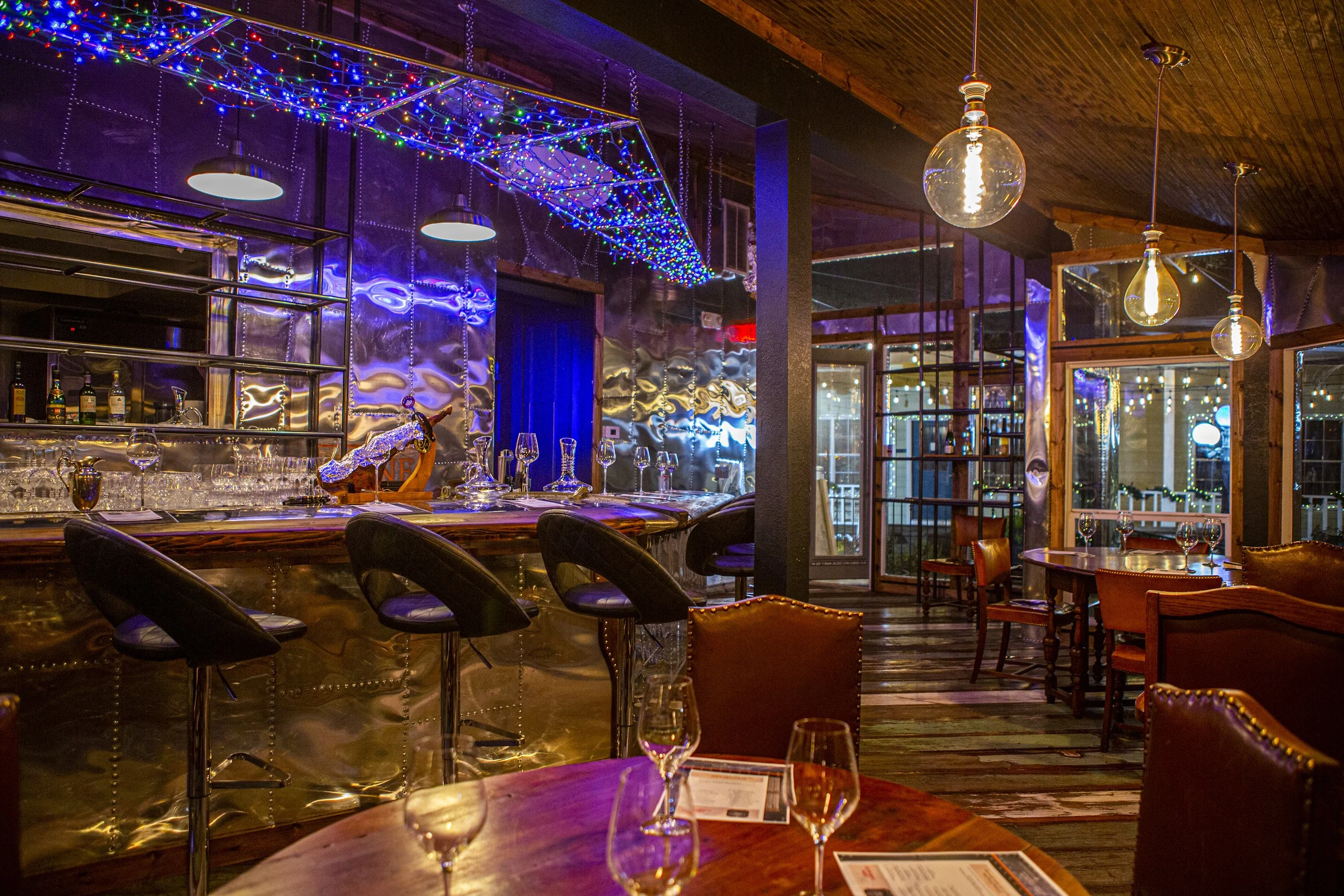 Interior of a restaurant or bar decorated for the holidays with colorful string lights and modern pendant lighting fixtures. There is a curved bar with upholstered chairs, and tables with wine glasses and menus on them. The space features a mix of wo