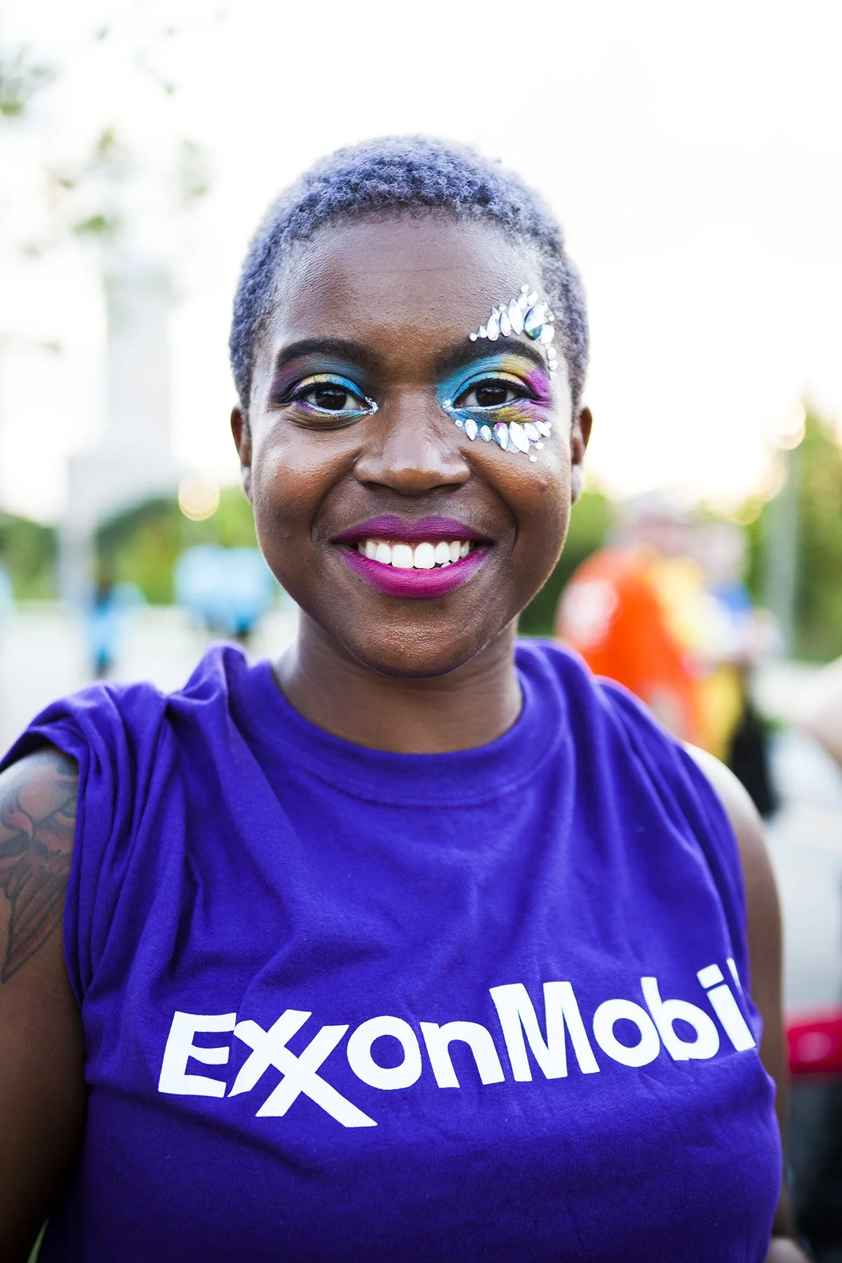 A woman with short, purple hair smiling, wearing a purple ExxonMobil T-shirt, and colorful face makeup with rhinestones near her eye.