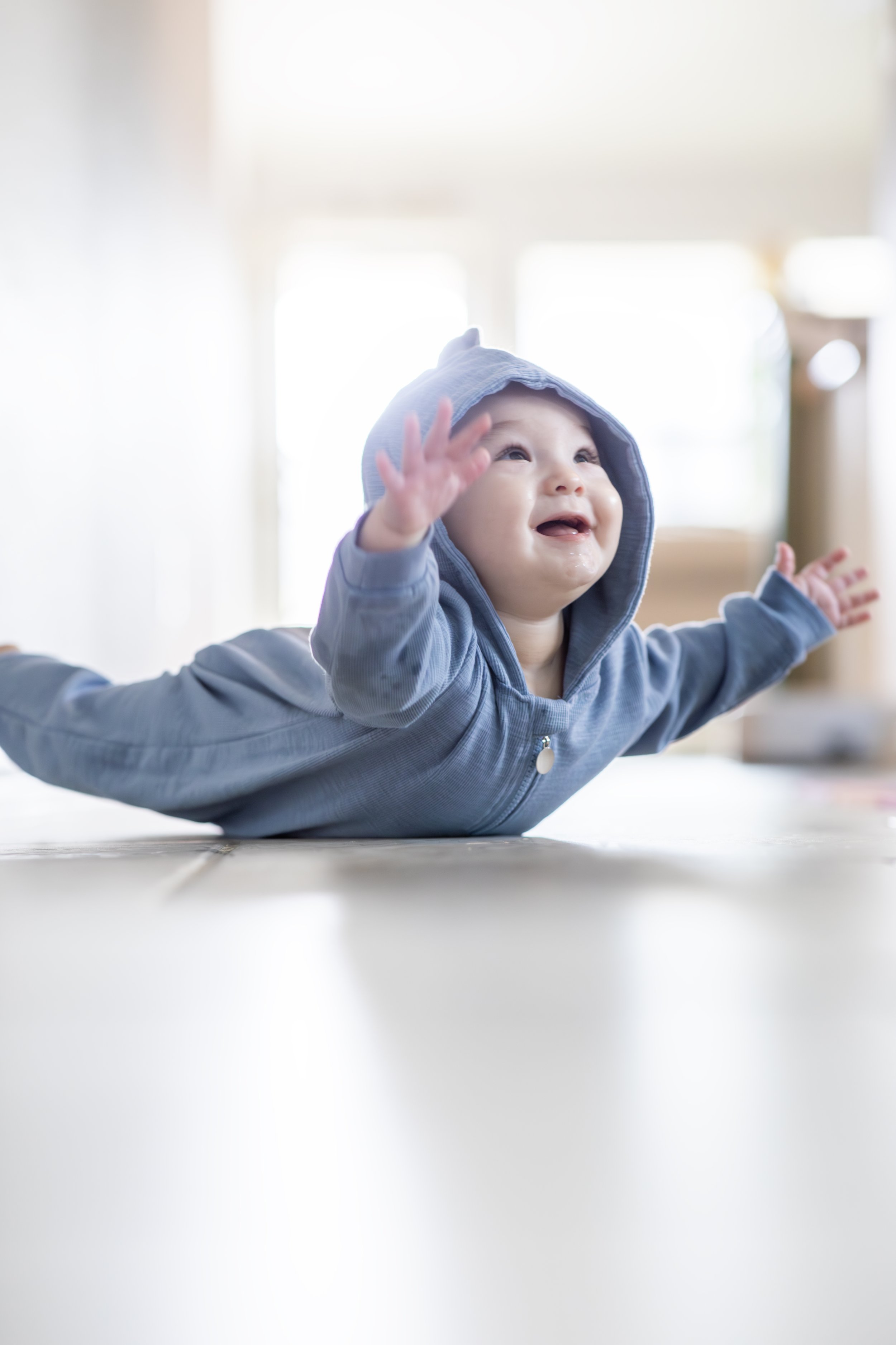 A young child in a blue hoodie lying on a white surface with arms outstretched and a happy expression. Photographed by Houston Photographer Katie Park.