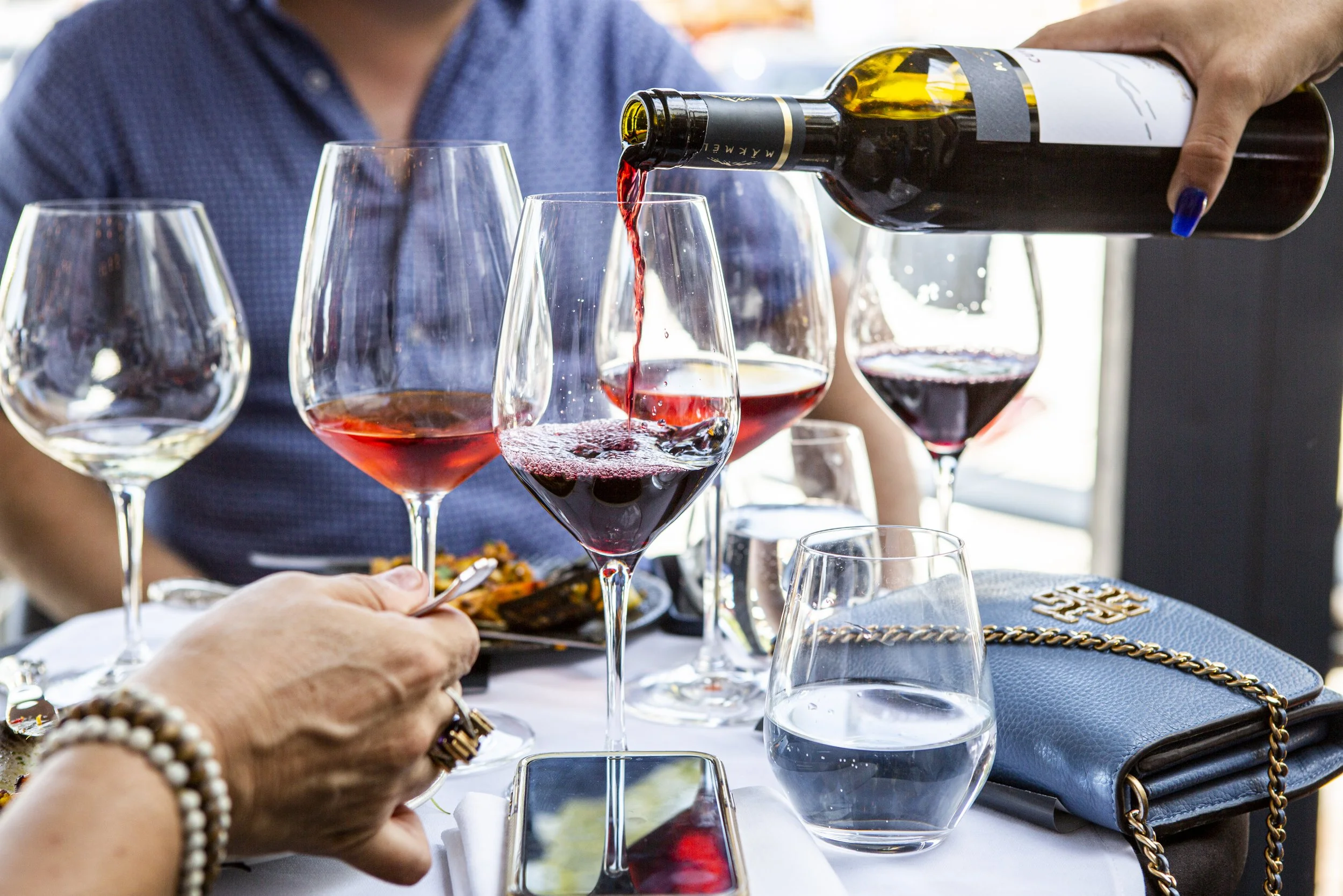 Person pouring red wine into a glass on a table with other wine glasses, a water glass, a blue purse with a chain strap, and a person in a blue shirt in the background.