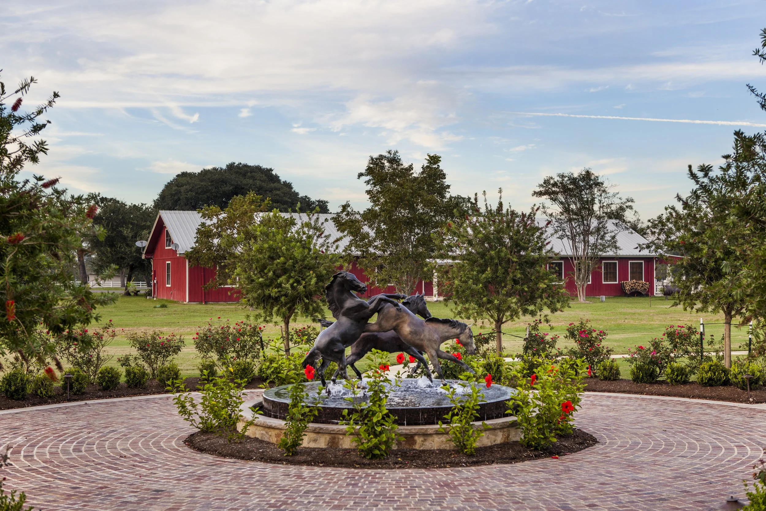A fountain with bronze horse statues in front of a red barn on a farm, surrounded by green trees, bushes, and a brick pathway during daytime.