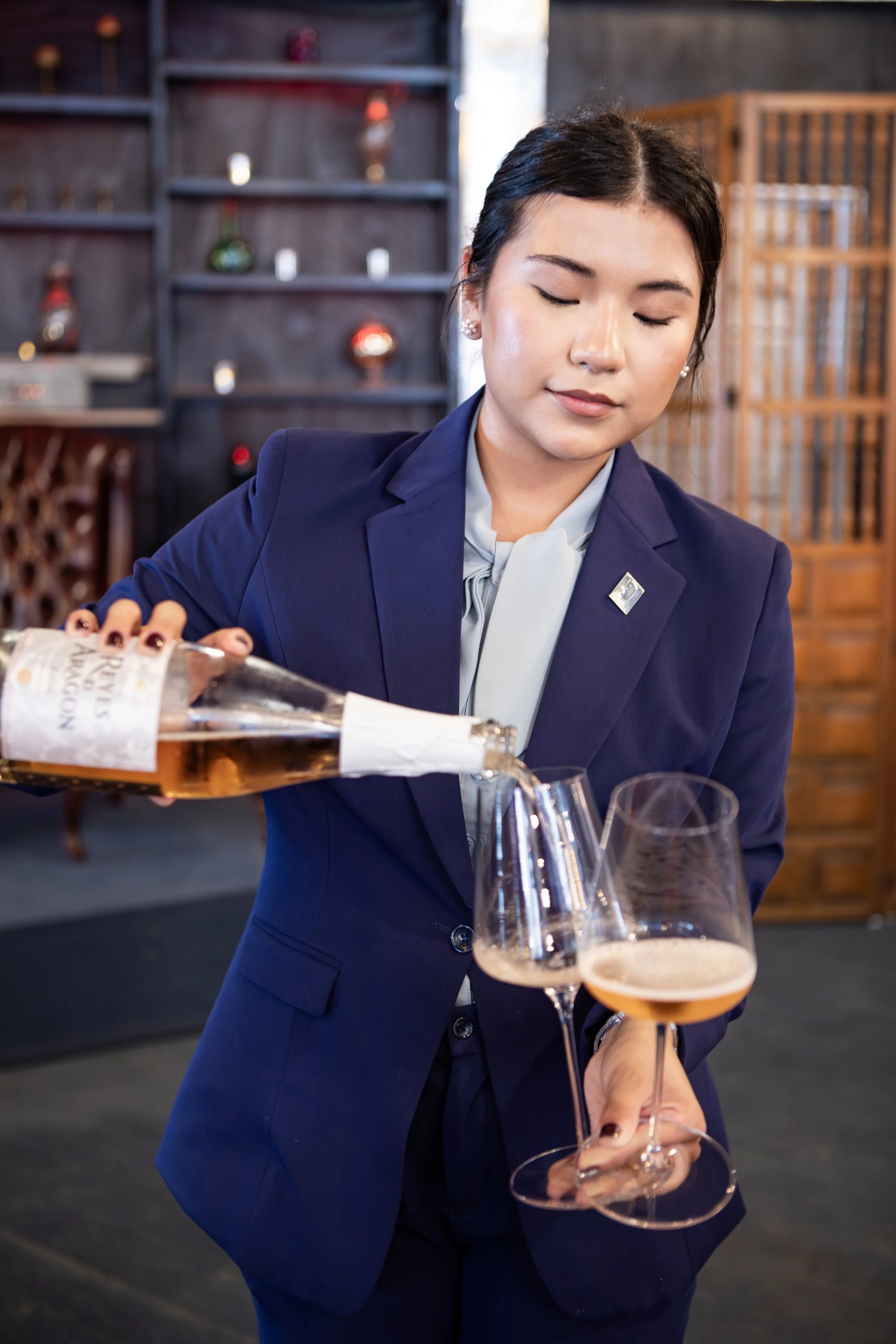 A woman in a navy blue suit pours wine into a glass in a cozy, stylish restaurant or bar setting.