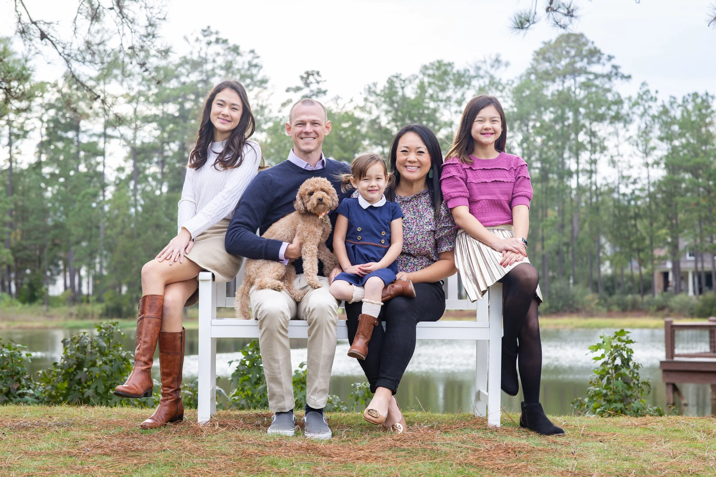Family of six, including two children and two adults, with a small dog, sitting on a white bench outdoors by a lake with trees in the background, smiling at the camera. Photographed by Houston Photographer Katie Park.