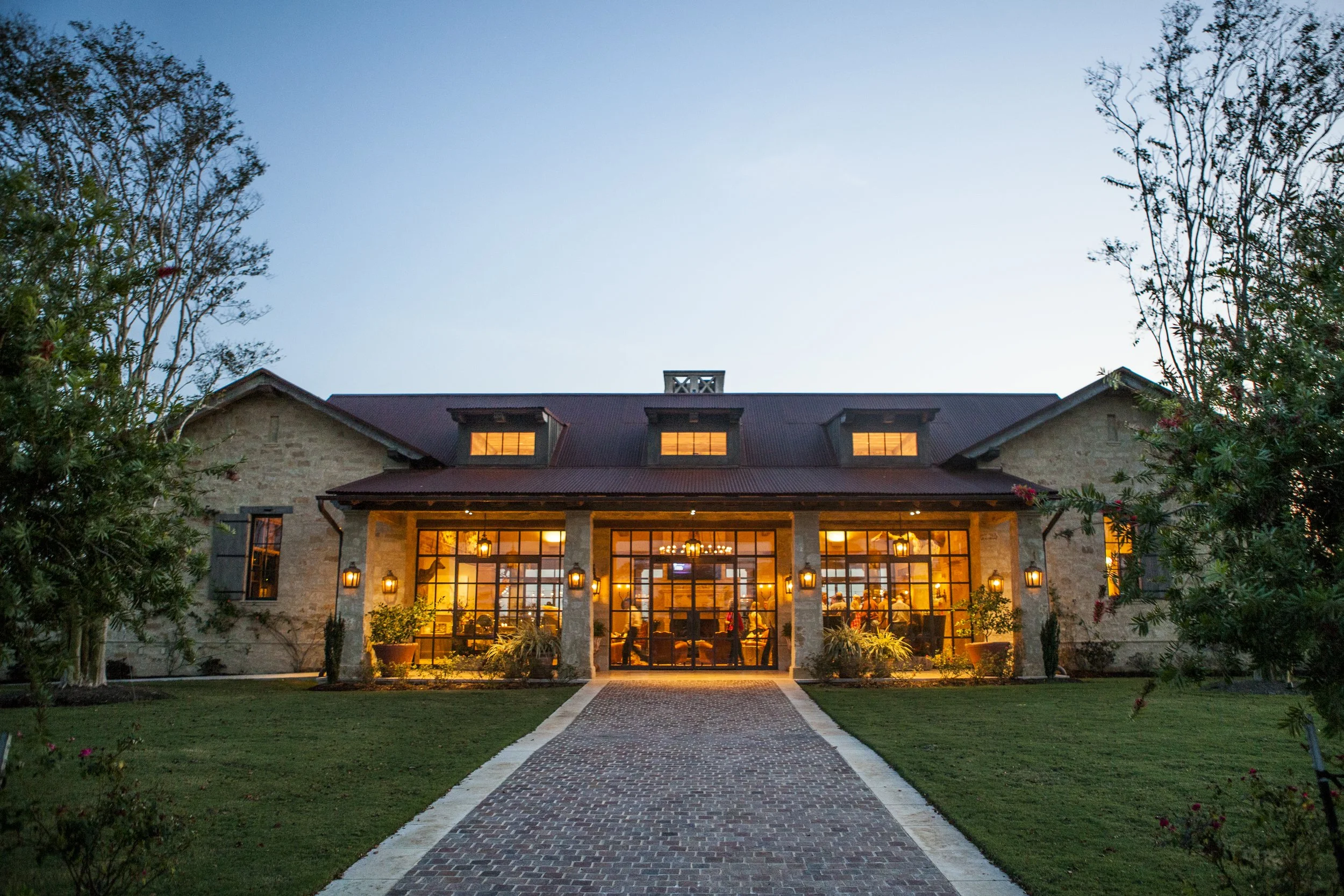 A large house with warm interior lighting, a brick pathway leading to the front entrance, surrounded by lush green lawn and trees, during dusk.