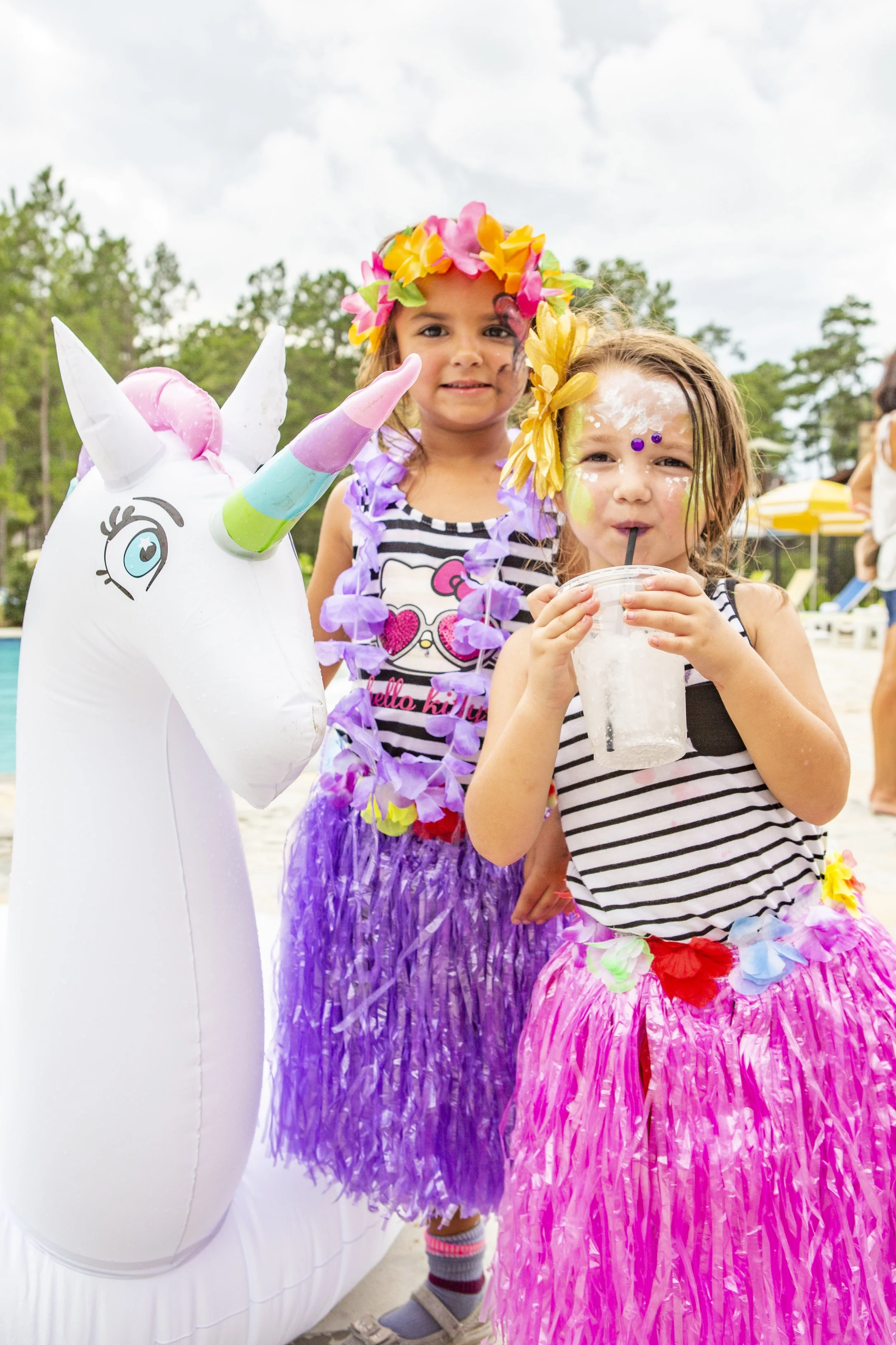 Two young girls dressed in colorful hula skirts and striped shirts, posing beside an inflatable unicorn, enjoying a festive outdoor event.