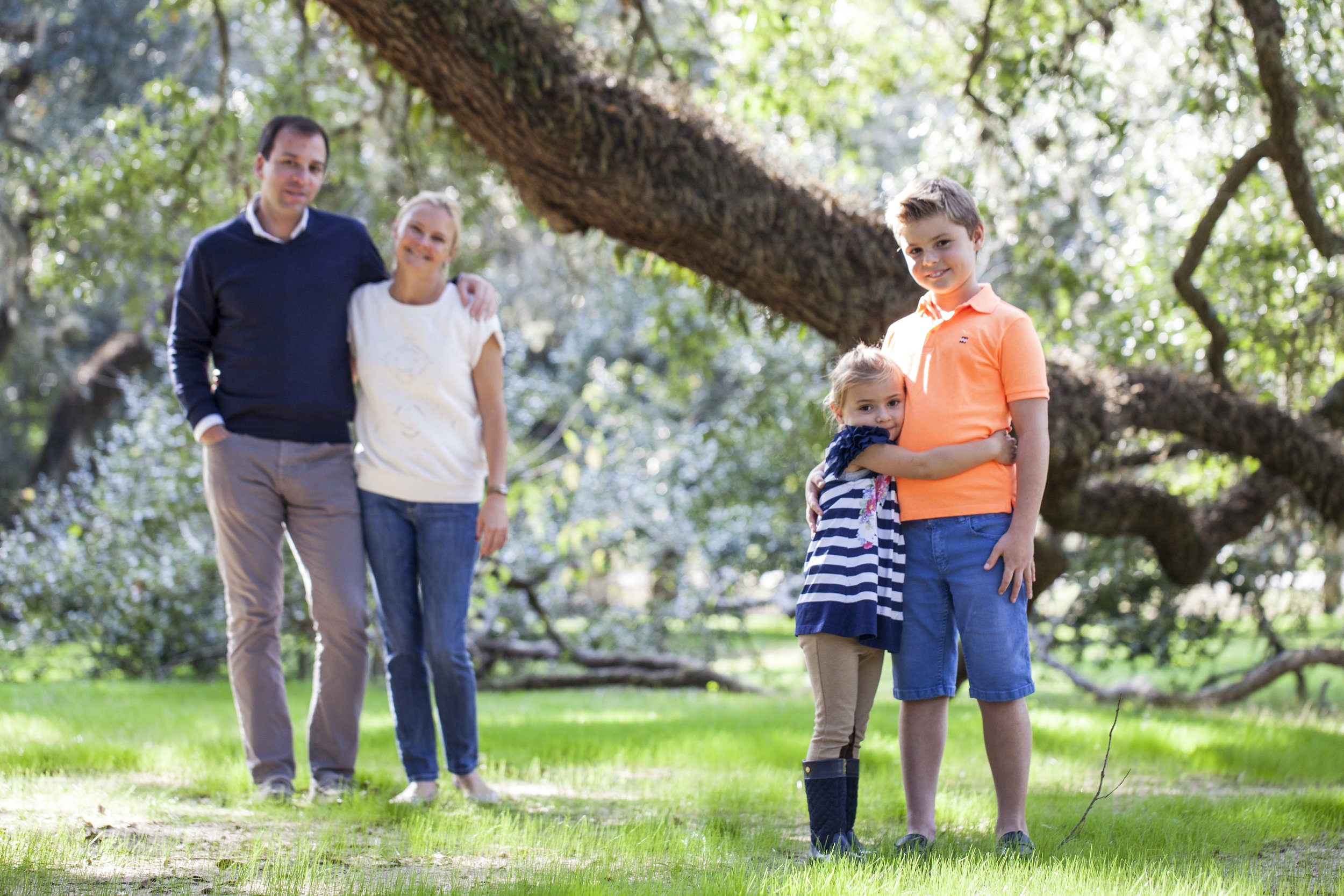 Family of four standing on grass in a park with large trees, smiling and hugging. Two children are in the foreground, a girl with a striped dress and a boy in an orange shirt, hugging each other. Behind them, a man and a woman stand close together, s