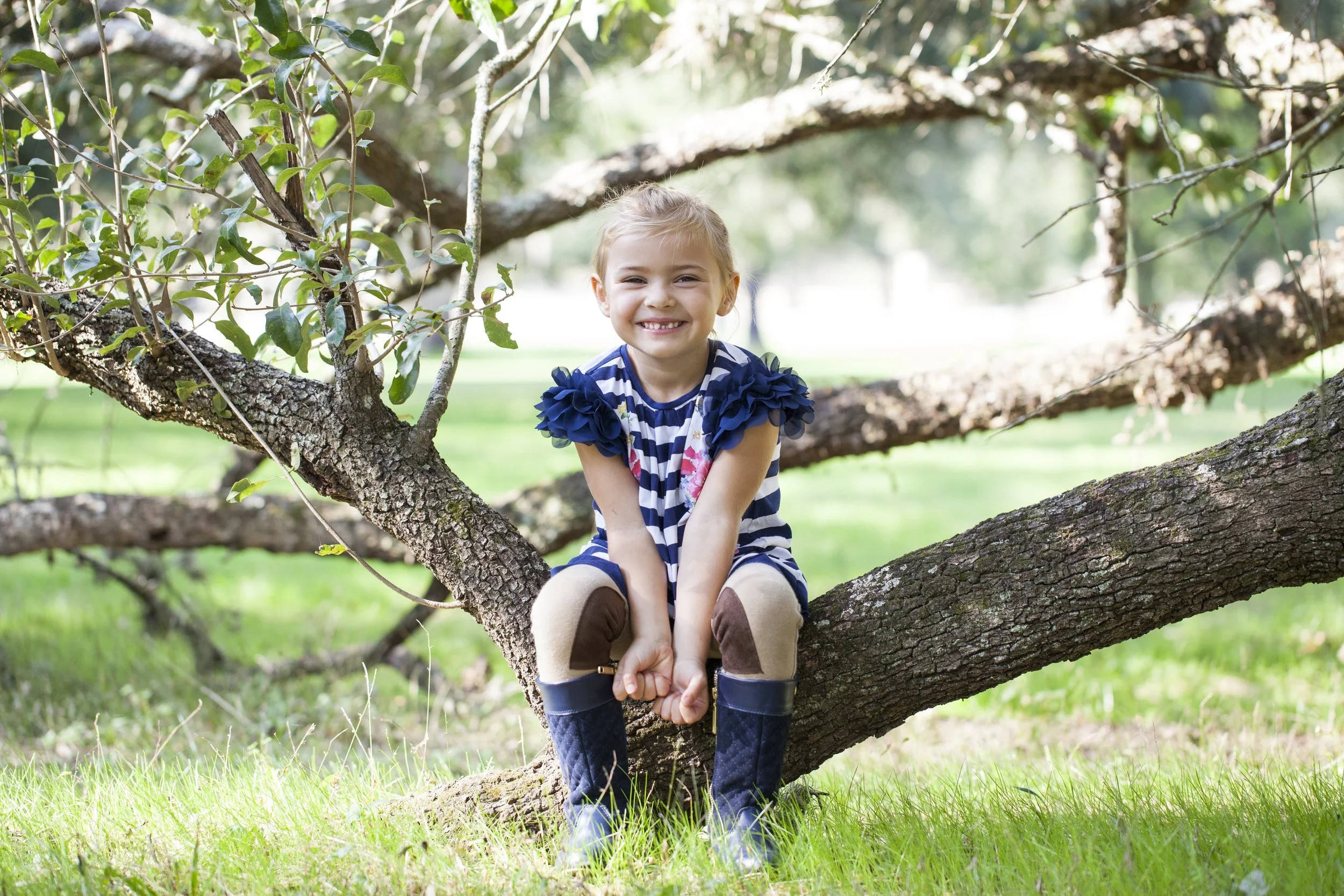 A young girl sitting on a tree branch outdoors, smiling at the camera, with green grass and trees in the background. Photographed by Houston Photographer Katie Park.