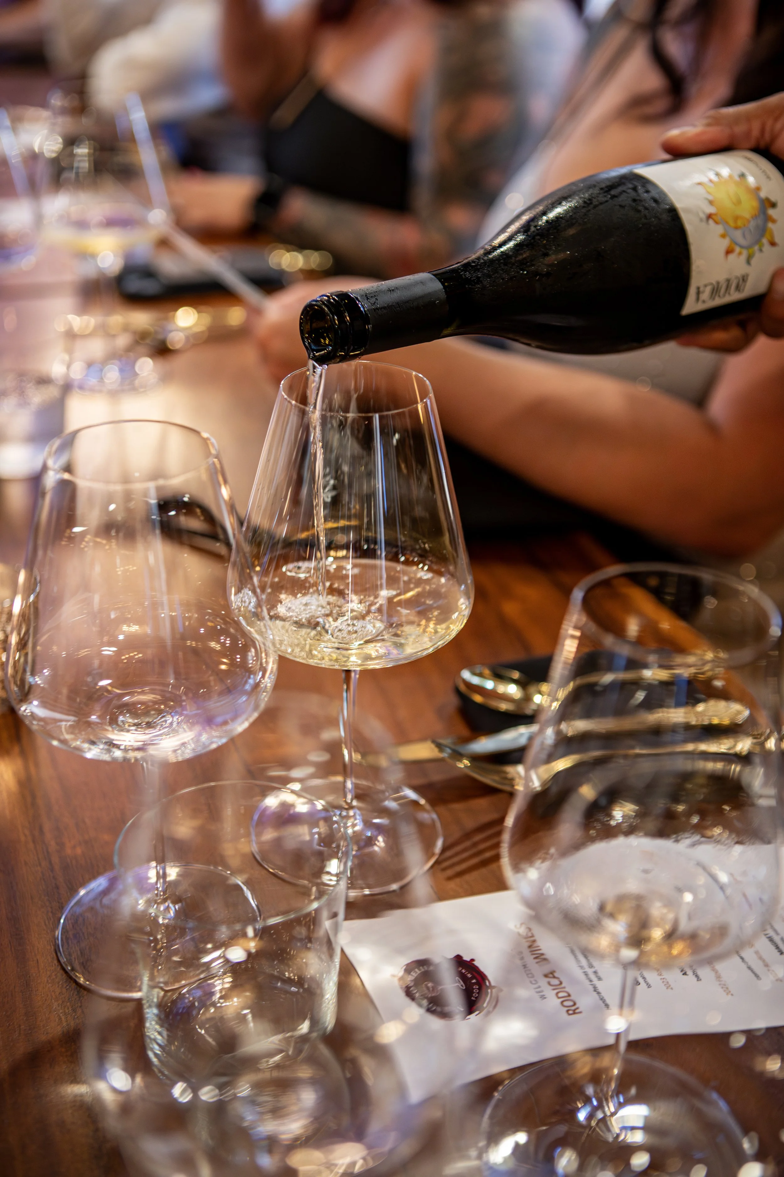 Person pouring white wine into a glass at a table with several empty wine glasses and a menu.