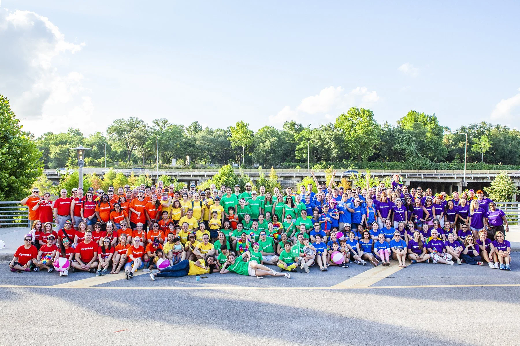 Group of people dressed in brightly colored shirts, posing outdoors on a clear day. The shirts are in red, orange, yellow, green, blue, and purple, and the group appears to be celebrating or participating in an event. They are seated and standing on 
