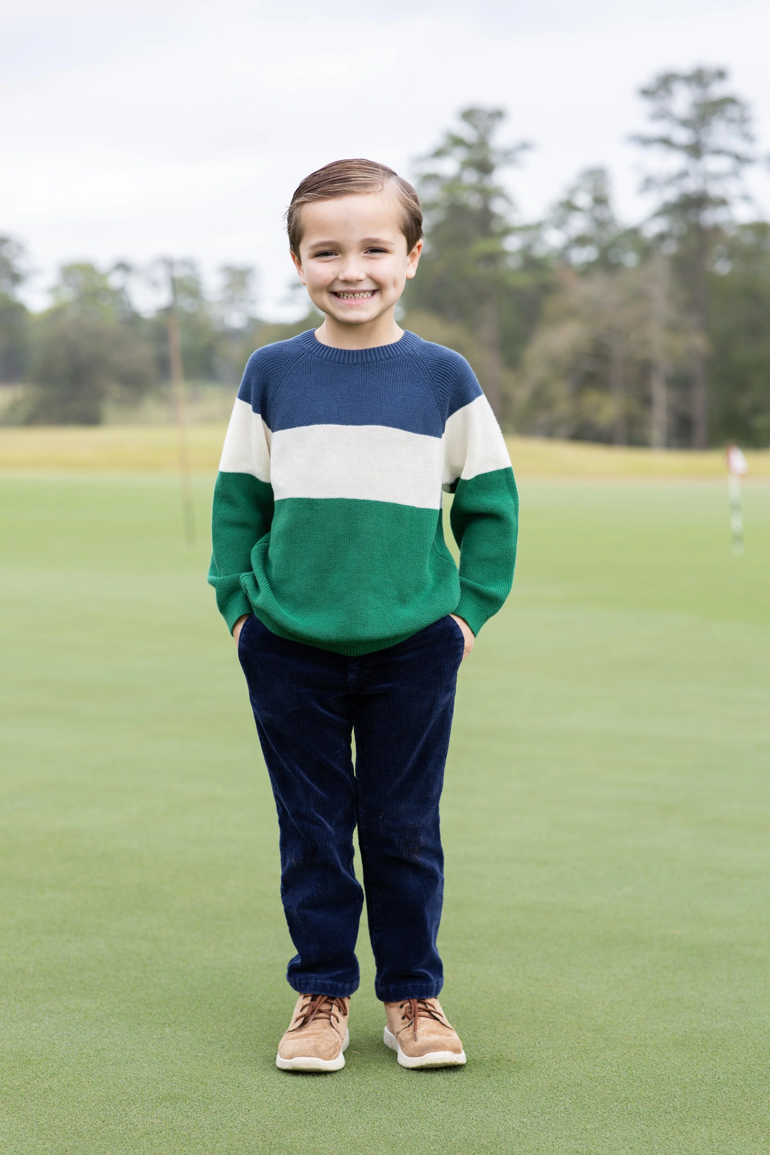 A young boy stands on a green golf course with his hands in his pockets, smiling at the camera, wearing a colorful striped sweater, dark pants, and tan shoes. Photographed by Houston Photographer Katie Park.