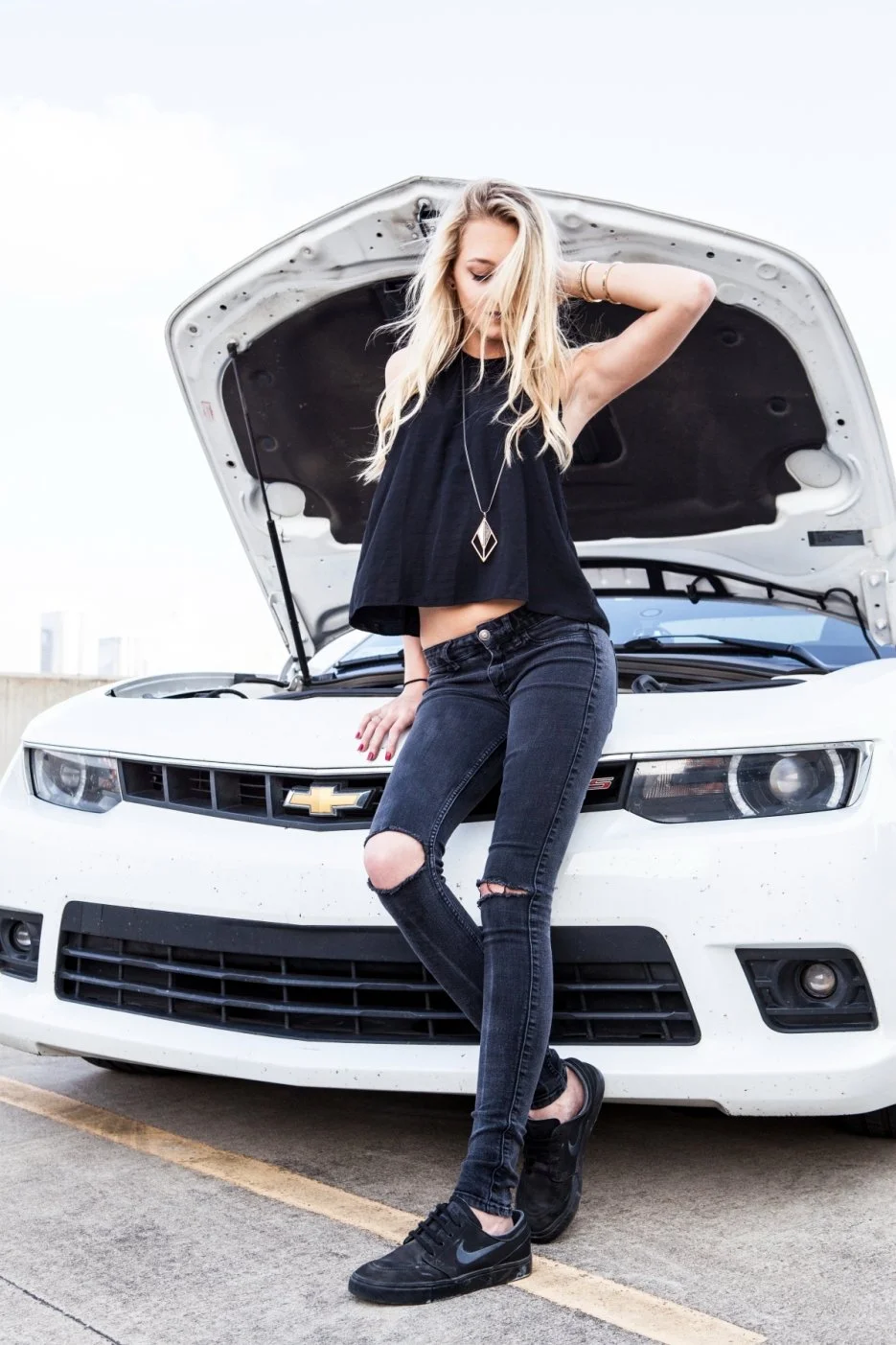 Young woman with blonde hair in a loose black top and ripped jeans standing in front of a white Chevrolet car with the hood open, on a parking lot during daytime. Photographed by Houston Photographer Katie Park.