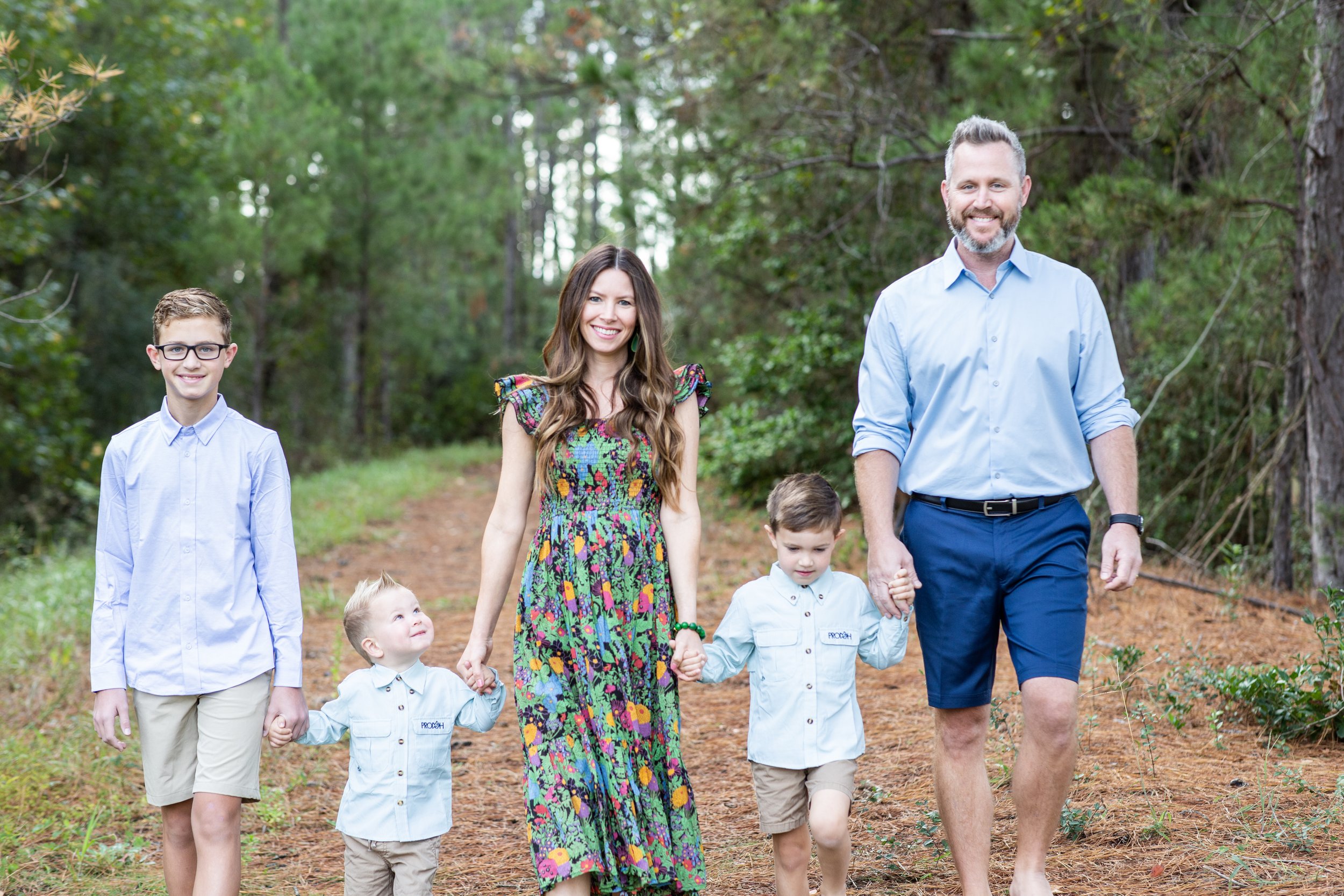 Family of six walking together outdoors on a trail in the woods, smiling, holding hands, dressed in casual summer clothes. Photographed by Houston Photographer Katie Park.