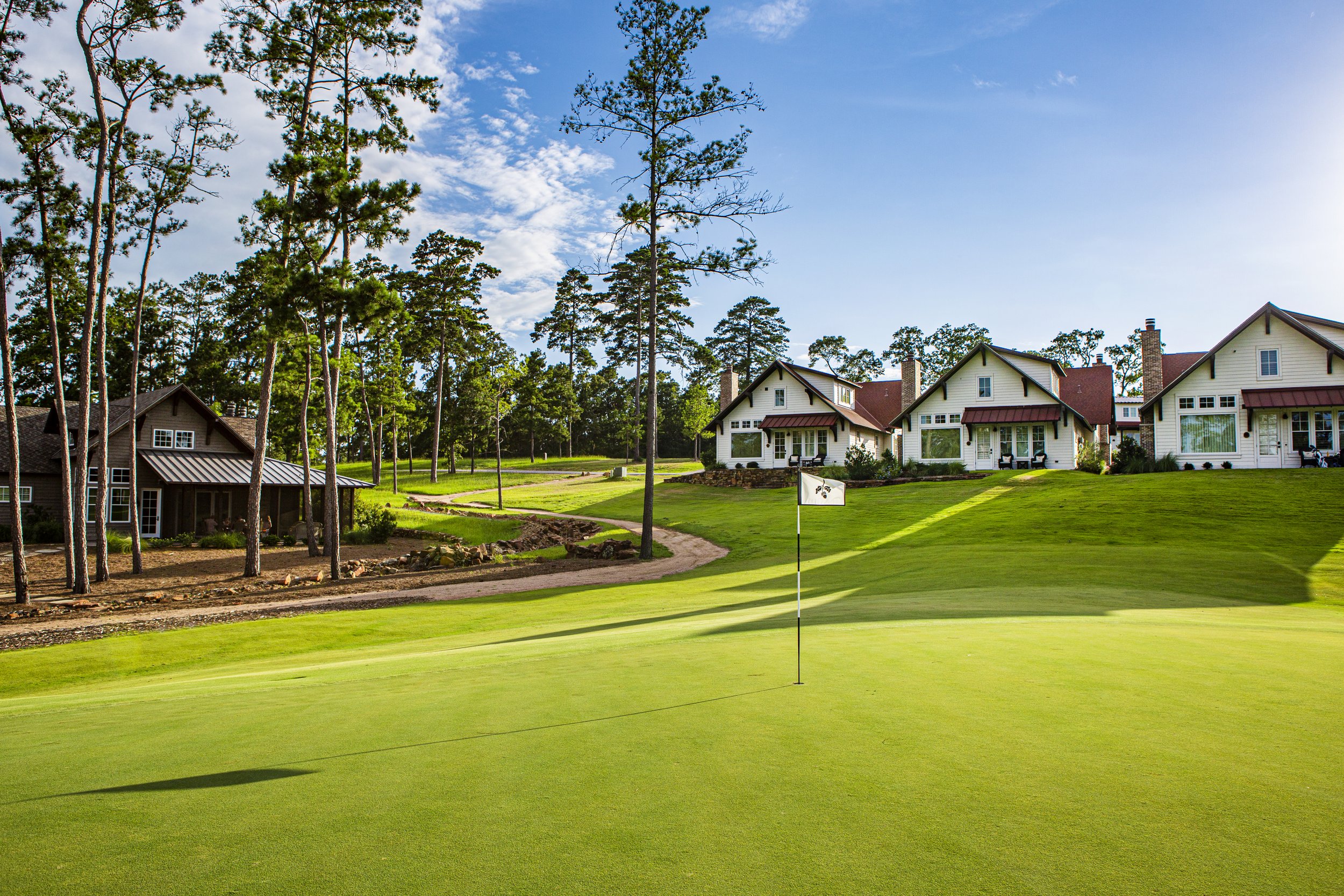 A golf course with a flag on the putting green, surrounded by residential houses and tall pine trees under a blue sky with scattered clouds.