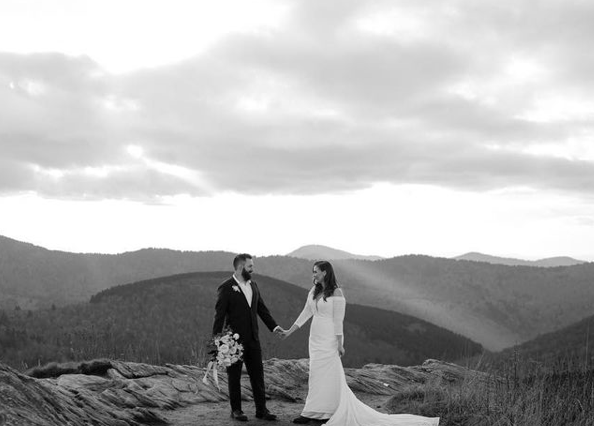 Black-and-white photo of a bride and groom holding hands on a rocky ledge with a mountain landscape in the background.