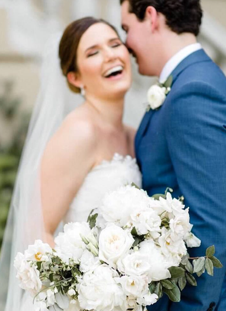A bride and groom celebrating their wedding, with the bride smiling and holding a large bouquet of white flowers, in front of a blurred outdoor background.