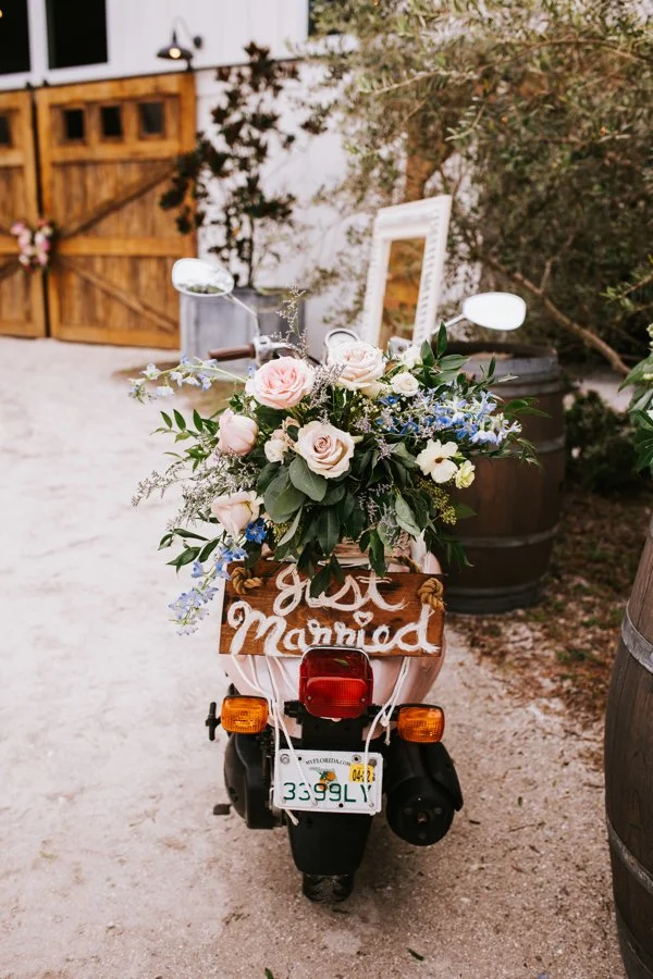 A motorcycle decorated with a large bouquet of pink and white roses, greenery, and other flowers, with a wooden sign reading "just married" attached to the back.