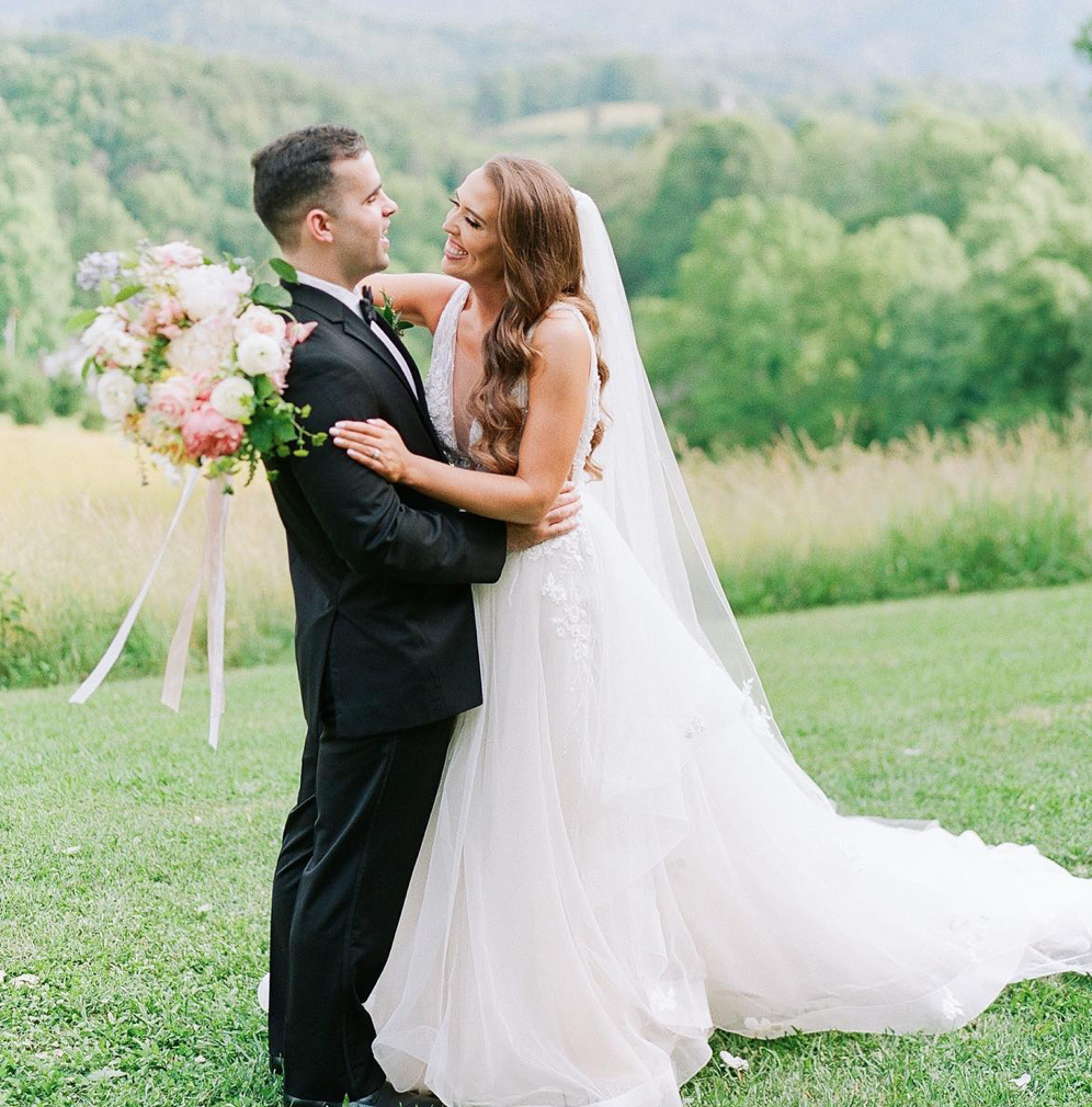 A bride and groom smiling and embracing outdoors on their wedding day, with the bride holding a bouquet of pink and white flowers, in a grassy field with trees in the background.
