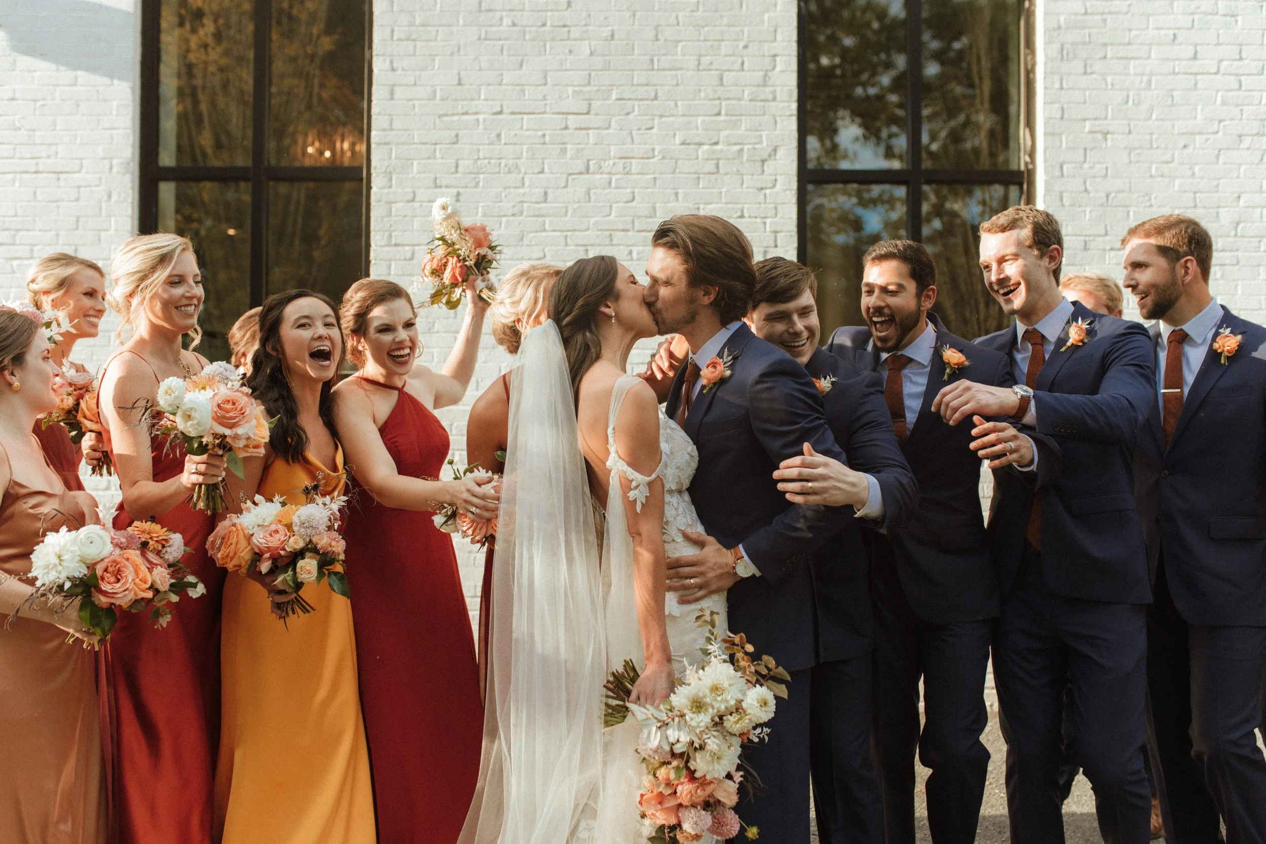 Bride and groom kissing surrounded by wedding party, with bridesmaids and groomsmen celebrating outdoors.