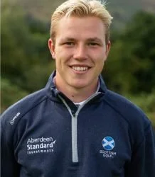 Man in blue jacket with logos, standing outdoors with a blurred natural background