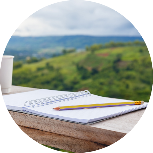 A journal, pen, and cup of coffee with a beautiful view of rolling hills in the background