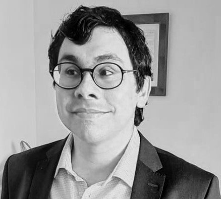 Homem com óculos de armação redonda, cabelo escuro e cabelo ondulado, vestindo blazer e camisa, sorrindo levemente em ambiente interno.