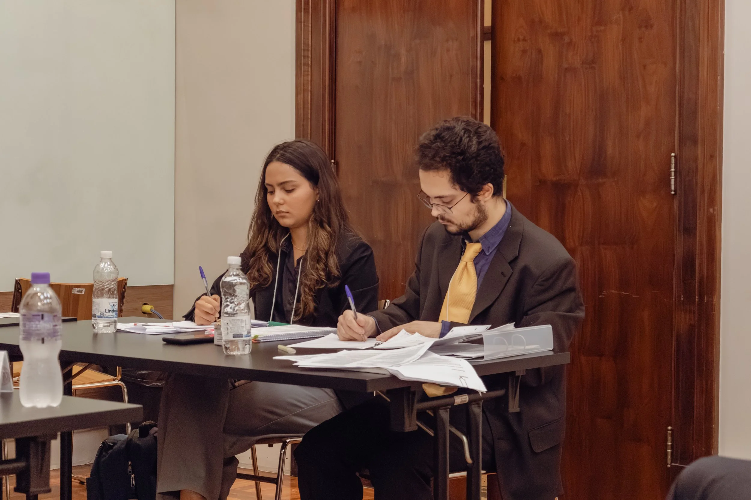 Duas pessoas sentadas em uma mesa de reunião, lendo e escrevendo em papéis, com várias garrafas de água na mesa, ambiente de sala de reunião ou auditório.
