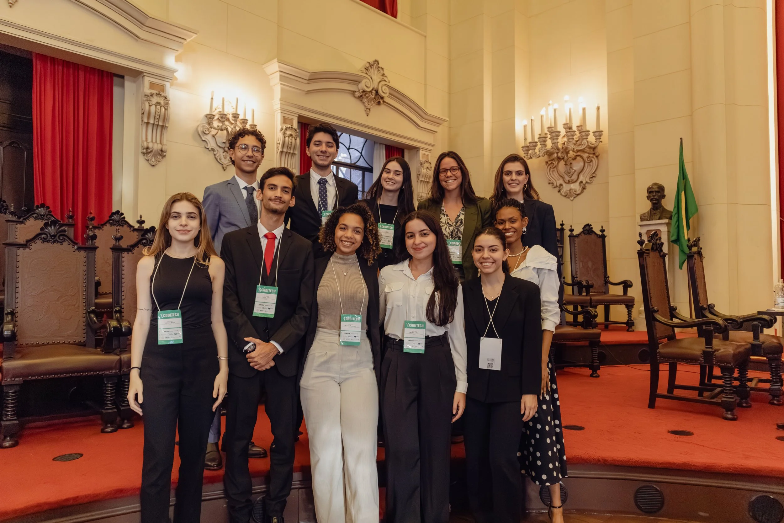 Grupo de jovens sorrindo em uma cerimônia formal, todos usando crachás, em um salão com decoração clássica e cadeiras de madeira no fundo.