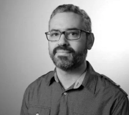 Homem com barba, cabelos grisalhos, usando óculos e camisa, retrato em preto e branco.