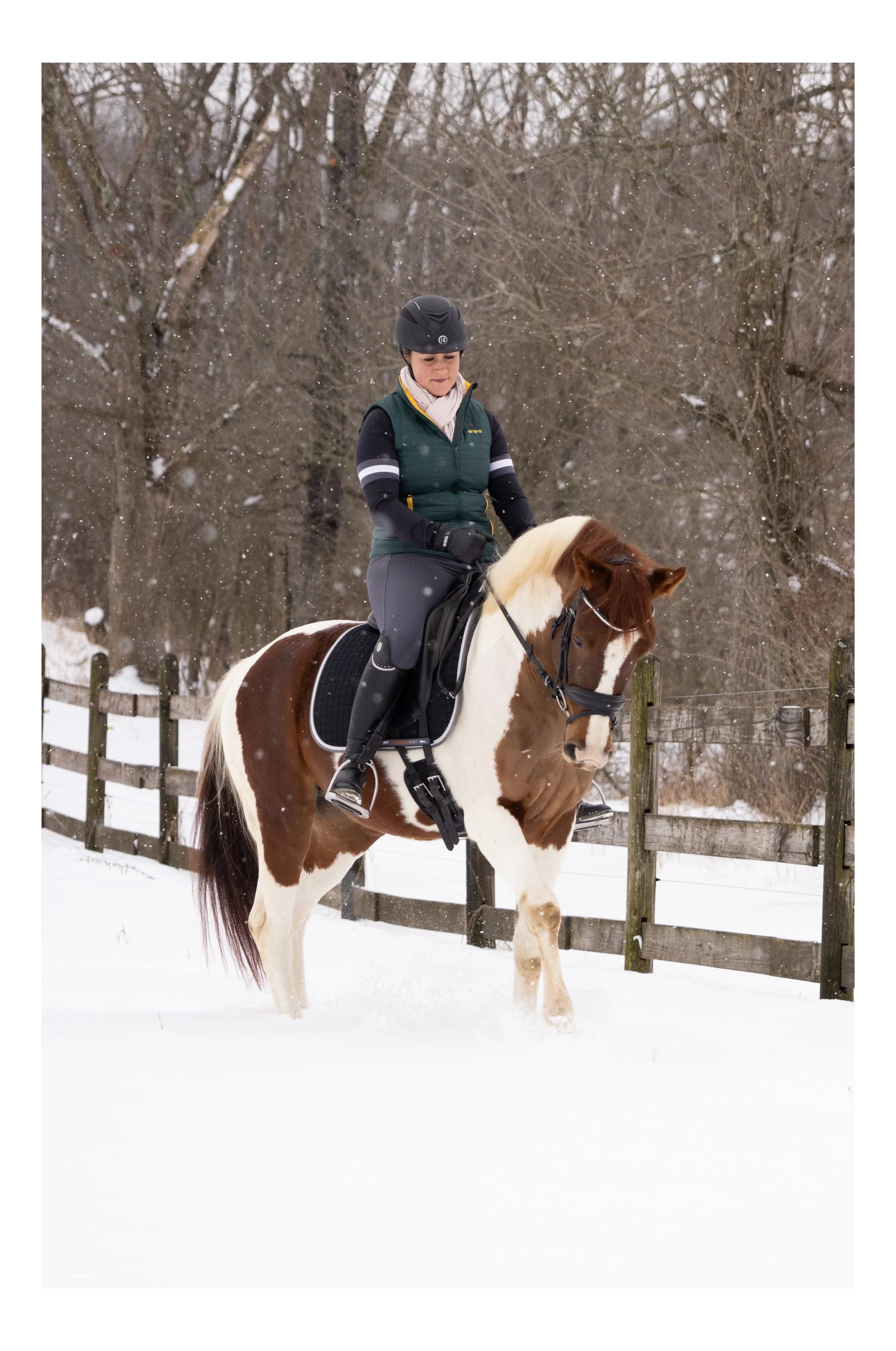 A woman wearing Ororo heated vest and a helmet riding a brown and white pinto horse through snow, with leafless trees and a wooden fence in the background.