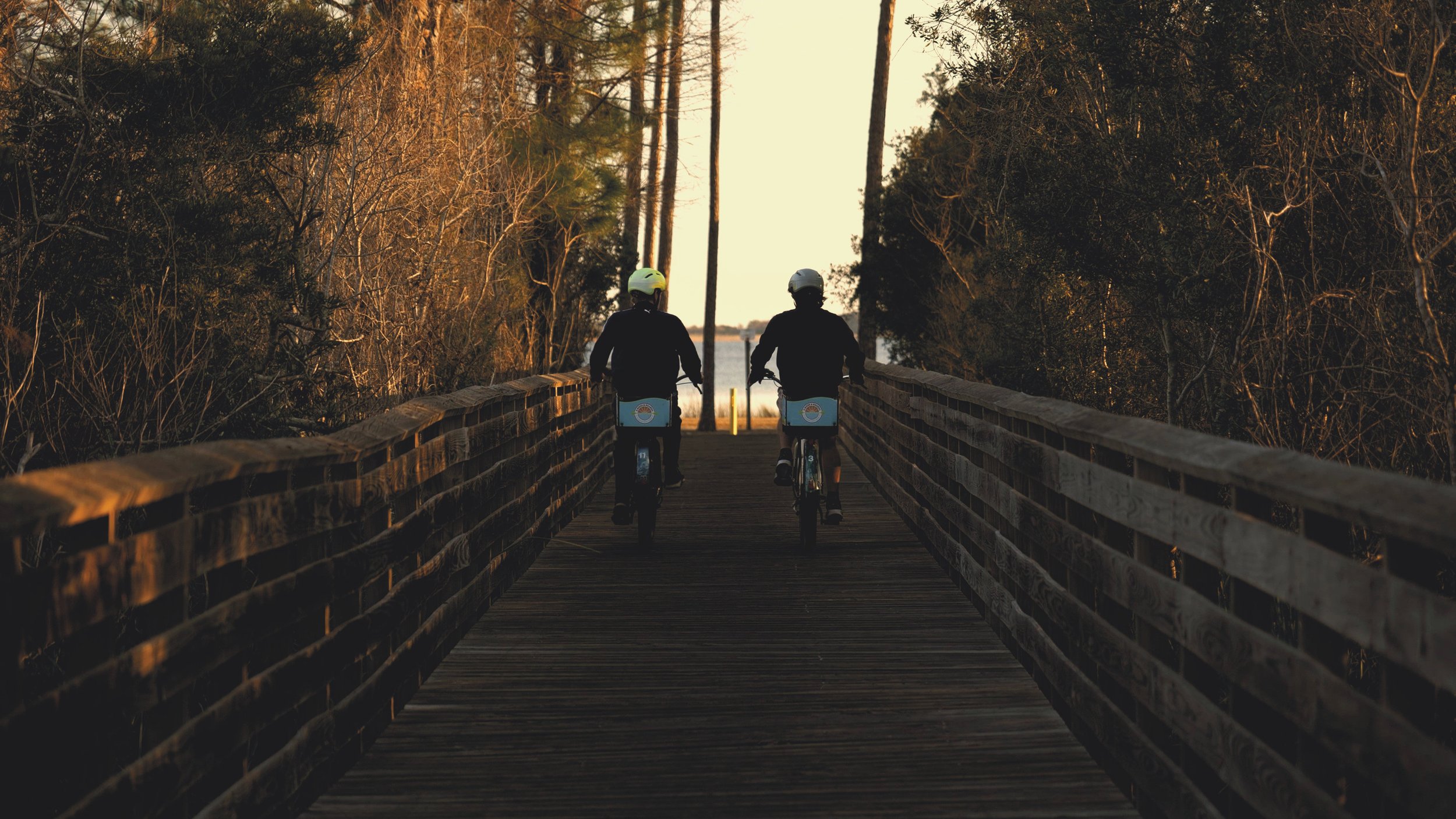 Two people riding e-bikes on a wooden bridge at Gulf State Park surrounded by trees, leading to a body of water in the distance during sunset.