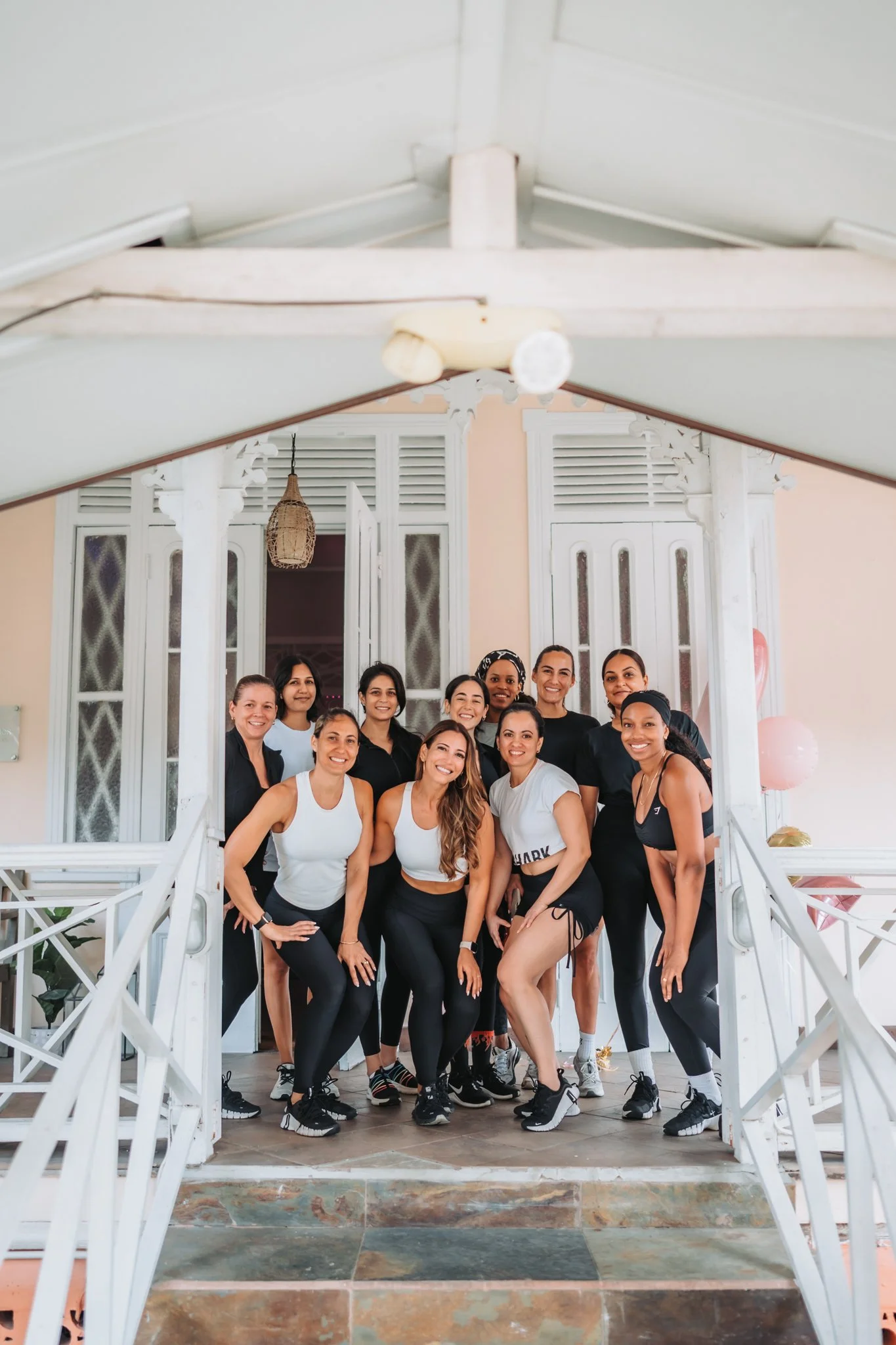 Group of women dressed in activewear posing on a porch with white decorative woodwork and open double doors in the background.
