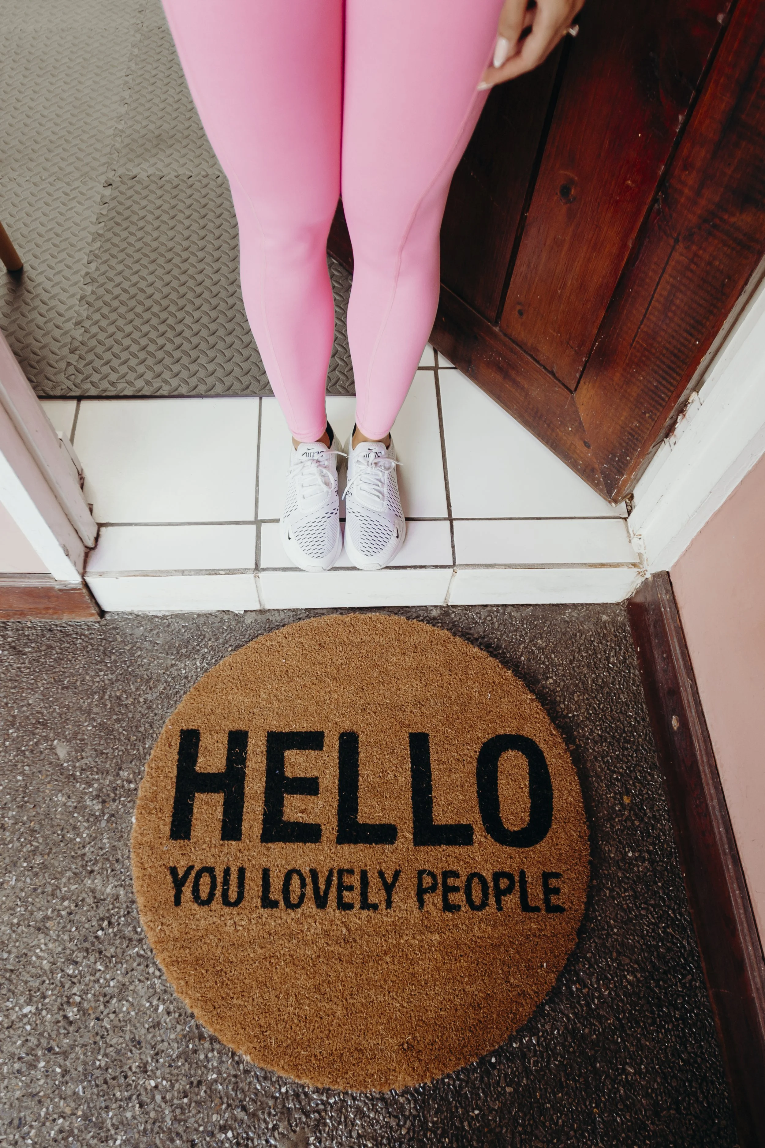 A person wearing pink leggings and white sneakers standing at the doorway, with a welcome mat that says, 'HELLO YOU LOVELY PEOPLE' in black letters.
