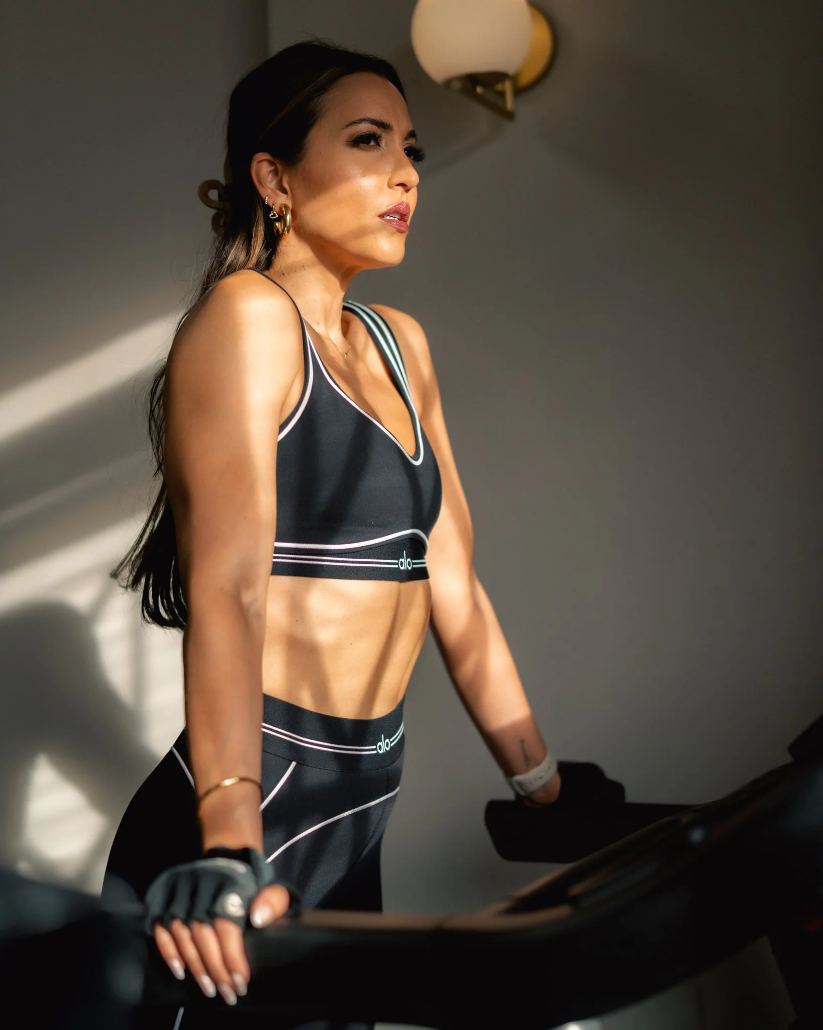 A woman in workout gear standing on a treadmill, looking to the side with sunlight on her face and a wall light in the background.