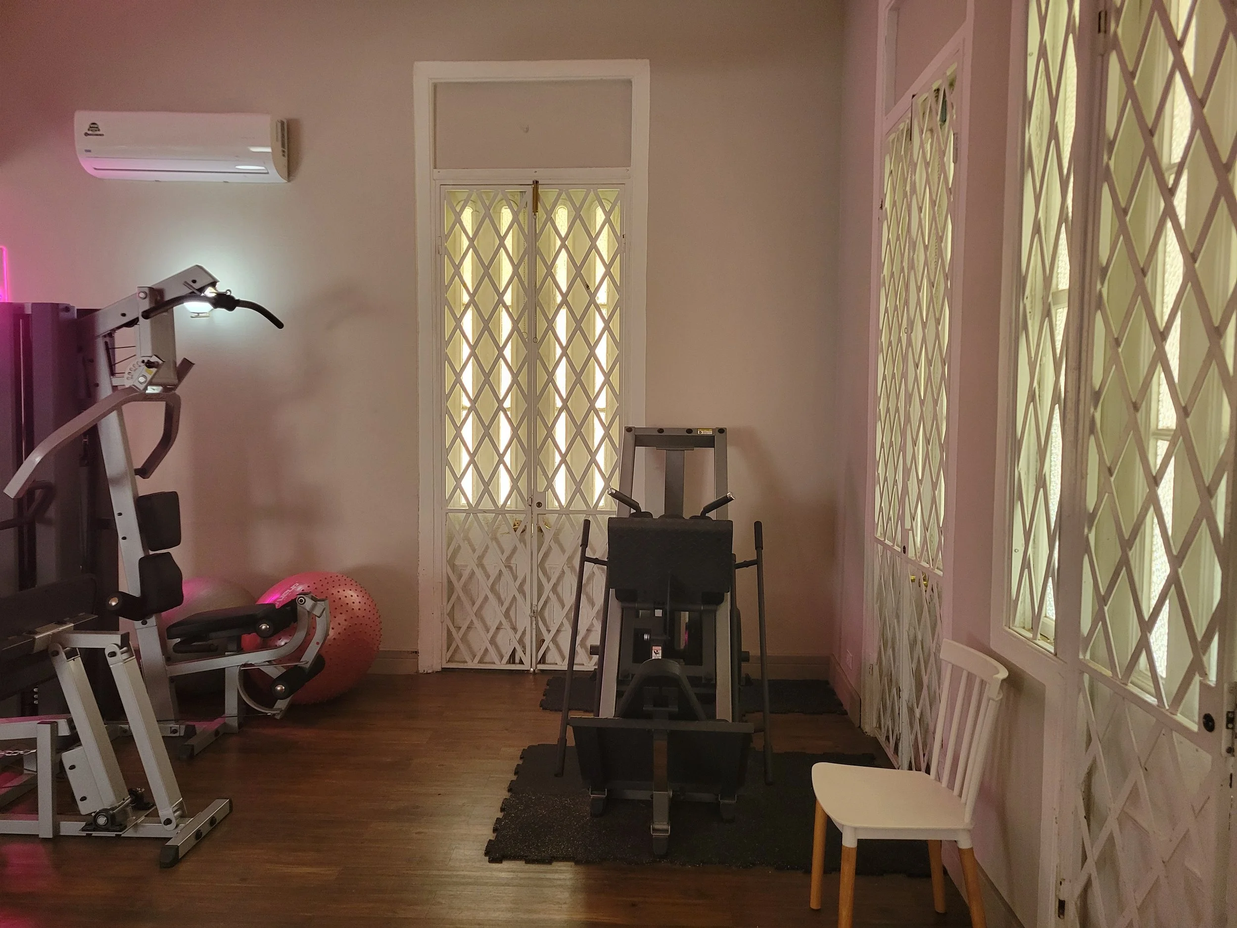 A workout room with exercise equipment, a chair, large window with lattice, an air conditioning unit, and pink lighting