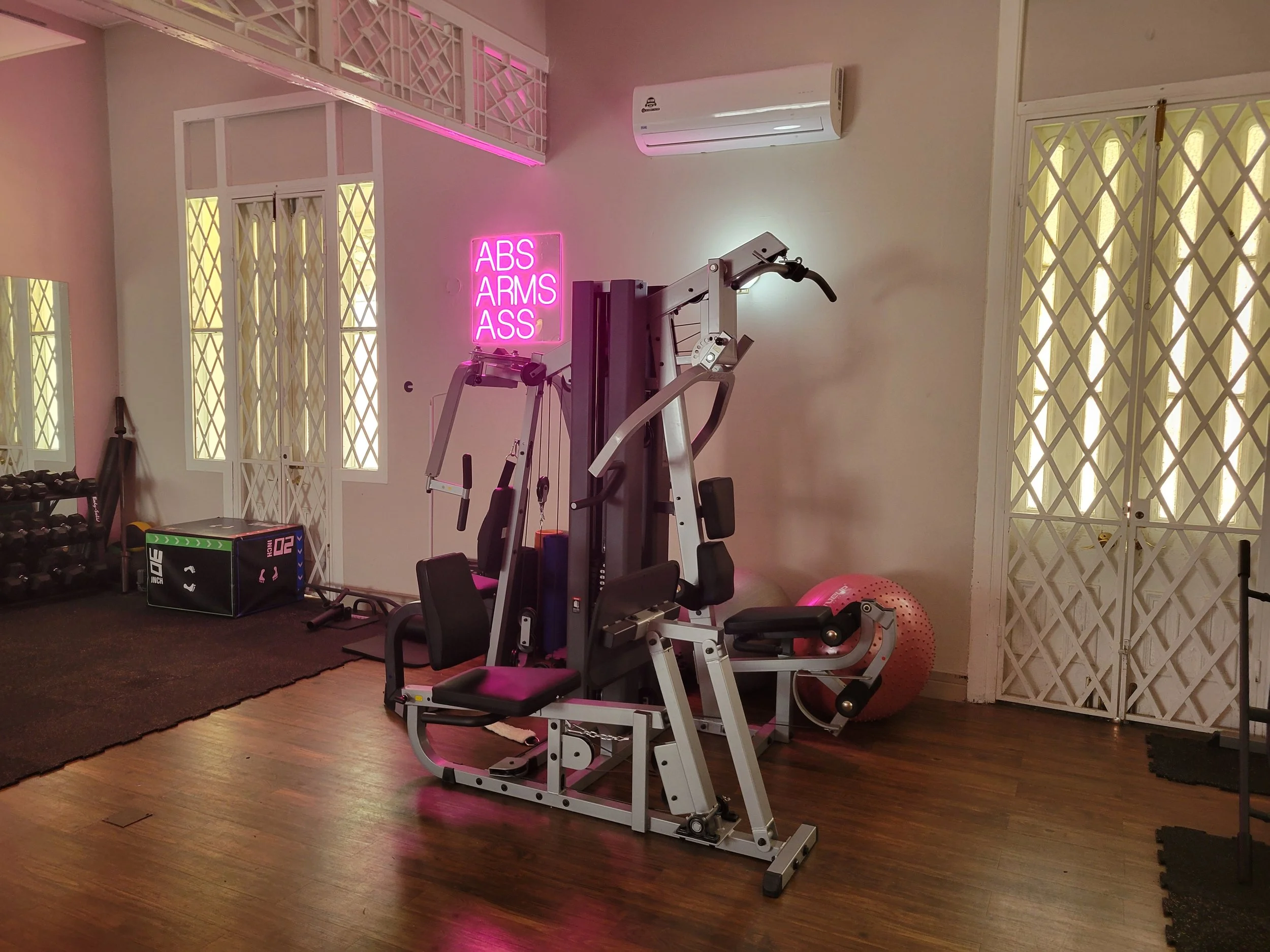 Home gym with workout equipment, including a weight machine, dumbbells, a pink exercise ball, and a neon pink sign that reads 'ABS ARMS ASS.' The room has wood flooring, white walls, and lattice-patterned doors and windows.