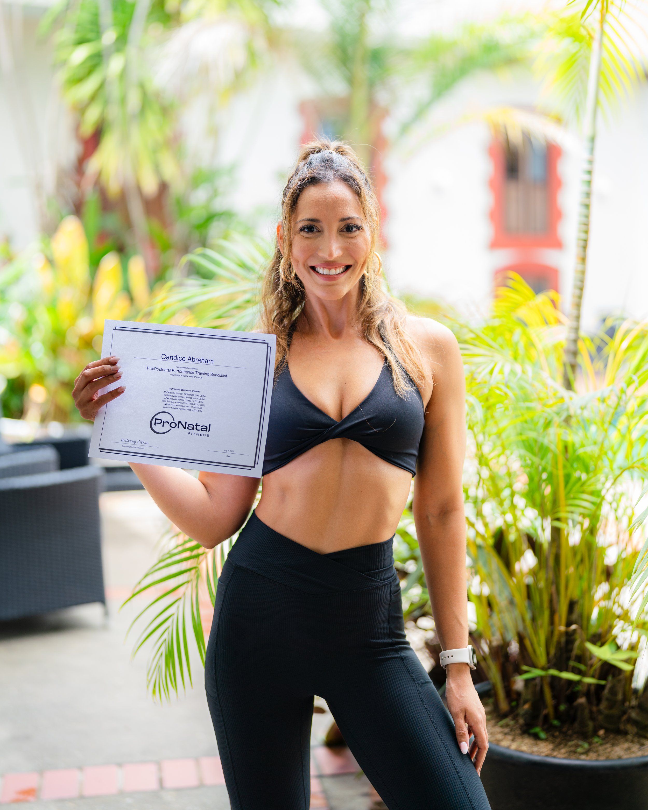 Smiling woman in workout attire holding a certification in an outdoor setting with plants.