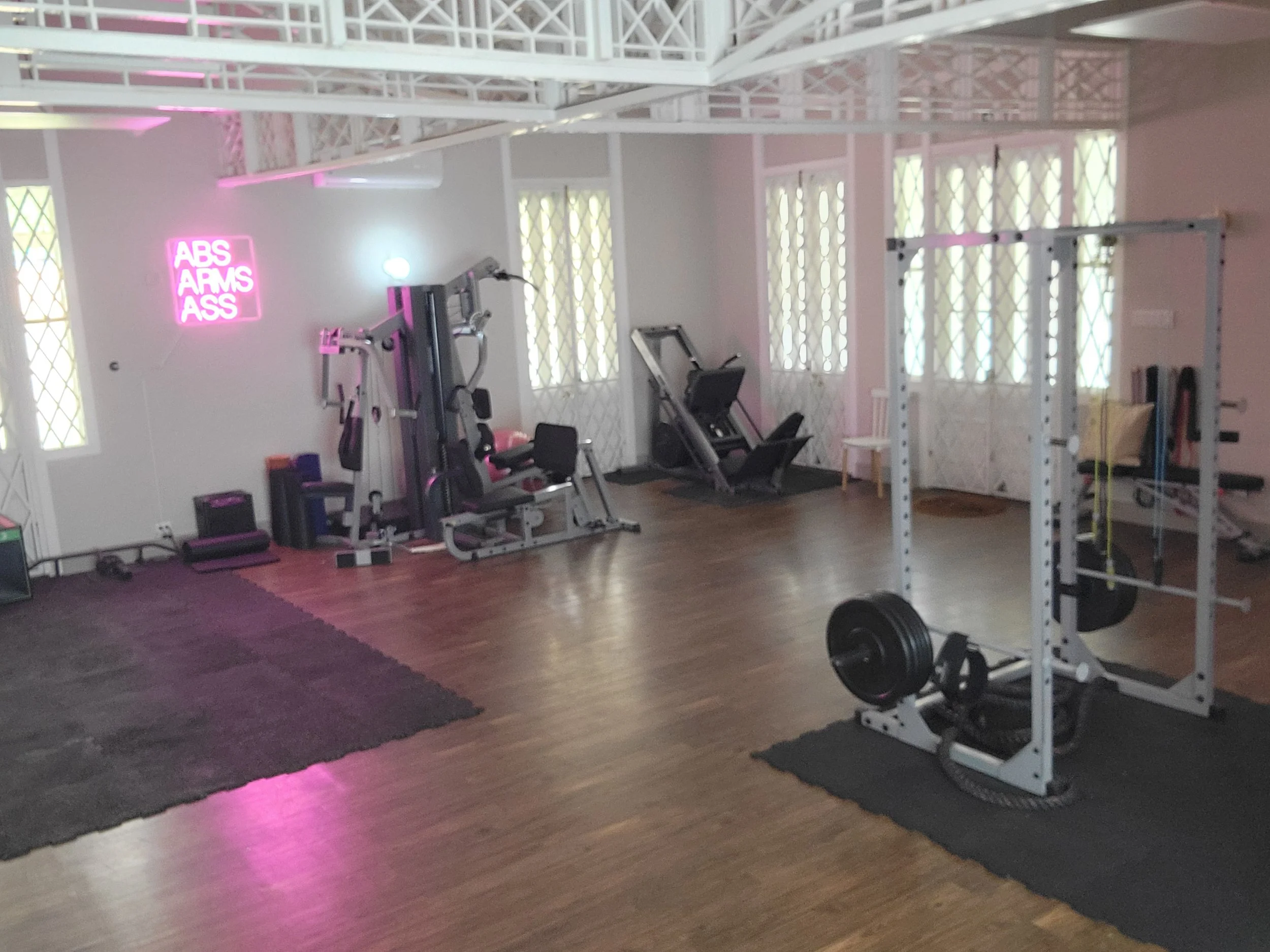 A spacious gym with various workout equipment, including a weight rack, exercise machines, and mats, illuminated by natural light from windows and a neon pink sign that reads 'ABS ARMS ASS'.