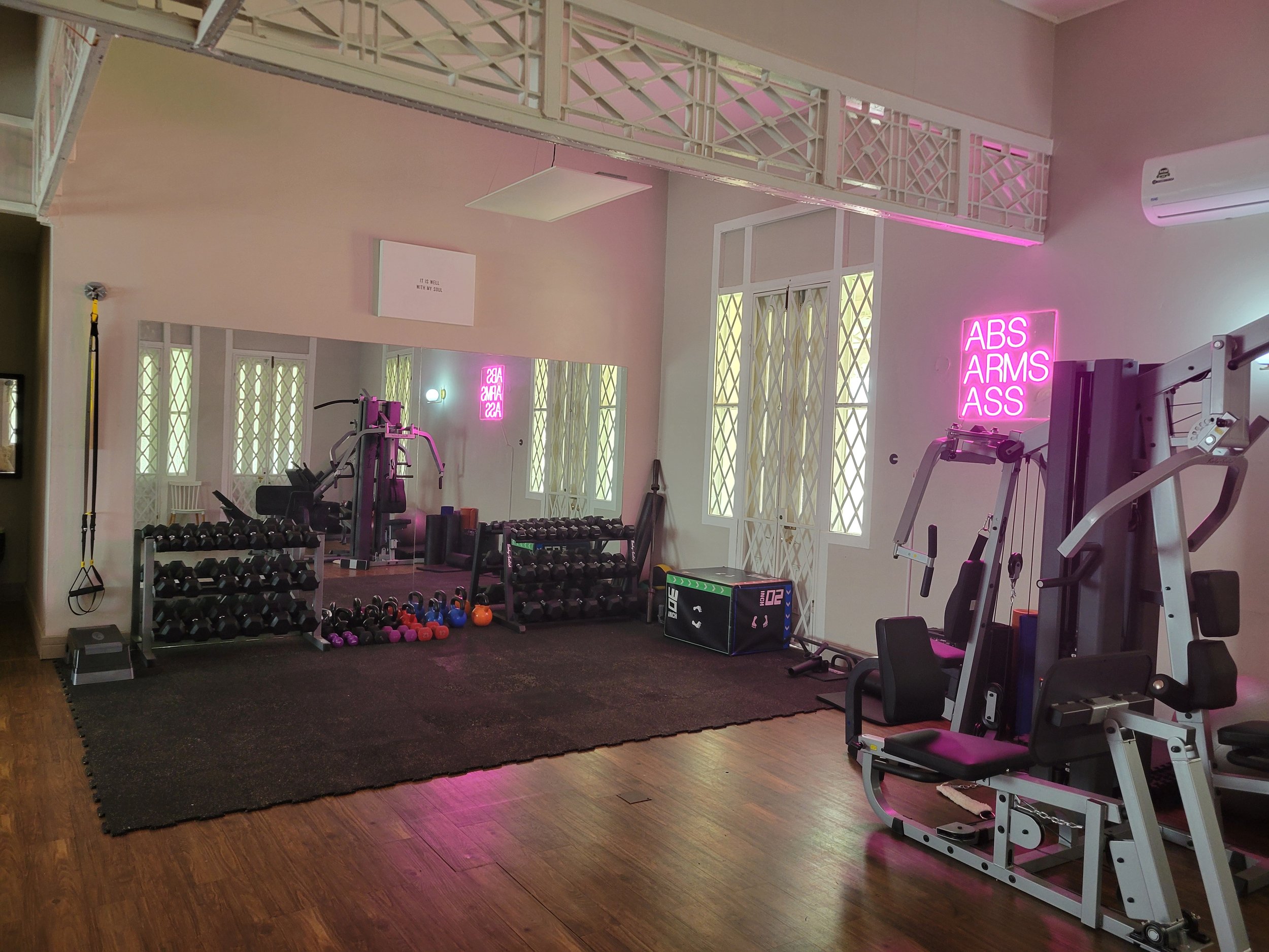 Interior of a gym with various workout equipment, including free weights, machines, and a kettlebell. Pink neon signs say 'ABS ARMS ASS' and 'GAS'. A large mirror reflects the room and windows with decorative grilles. The flooring is wood and rubber 