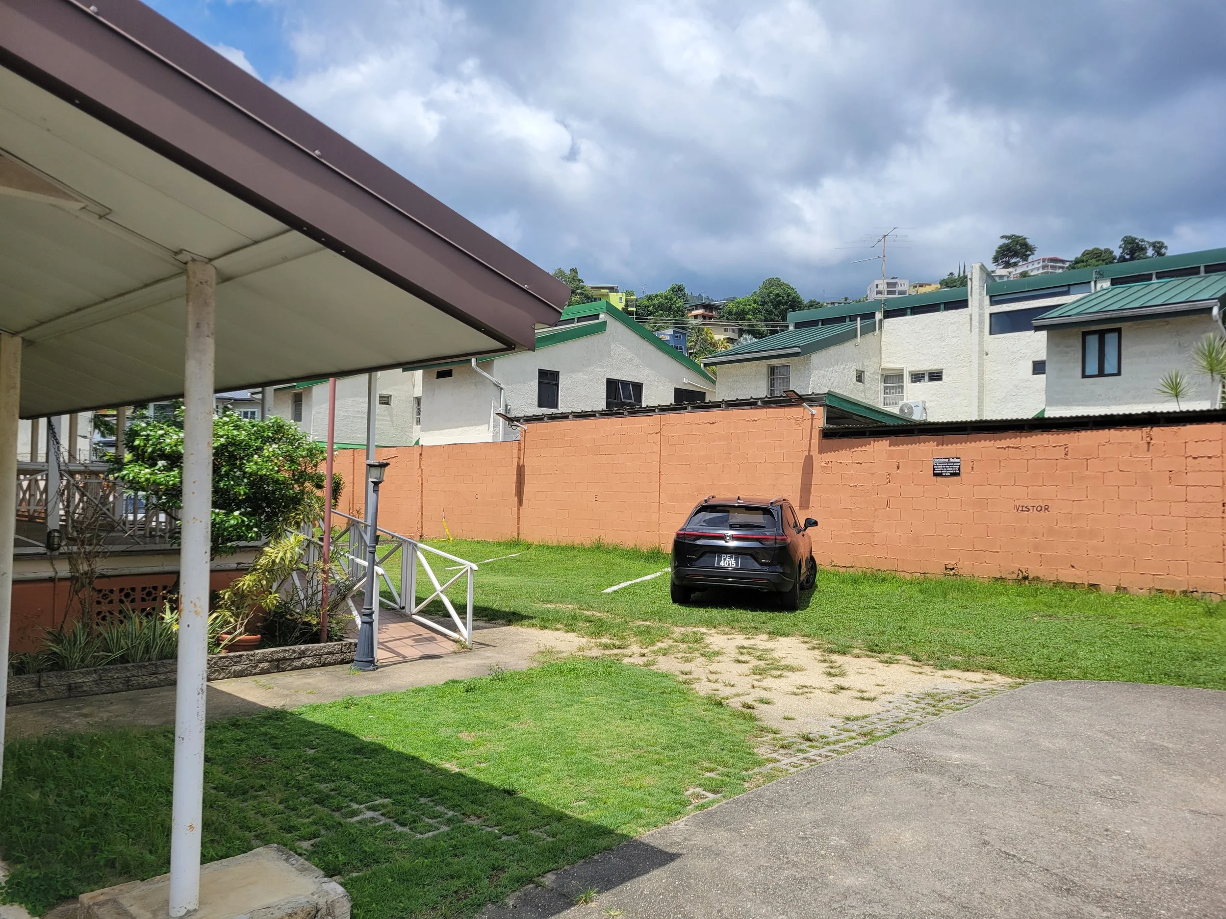 A parking lot with a black car parked on grass, a brick wall, residential buildings with green roofs, a sidewalk, and cloudy sky above.