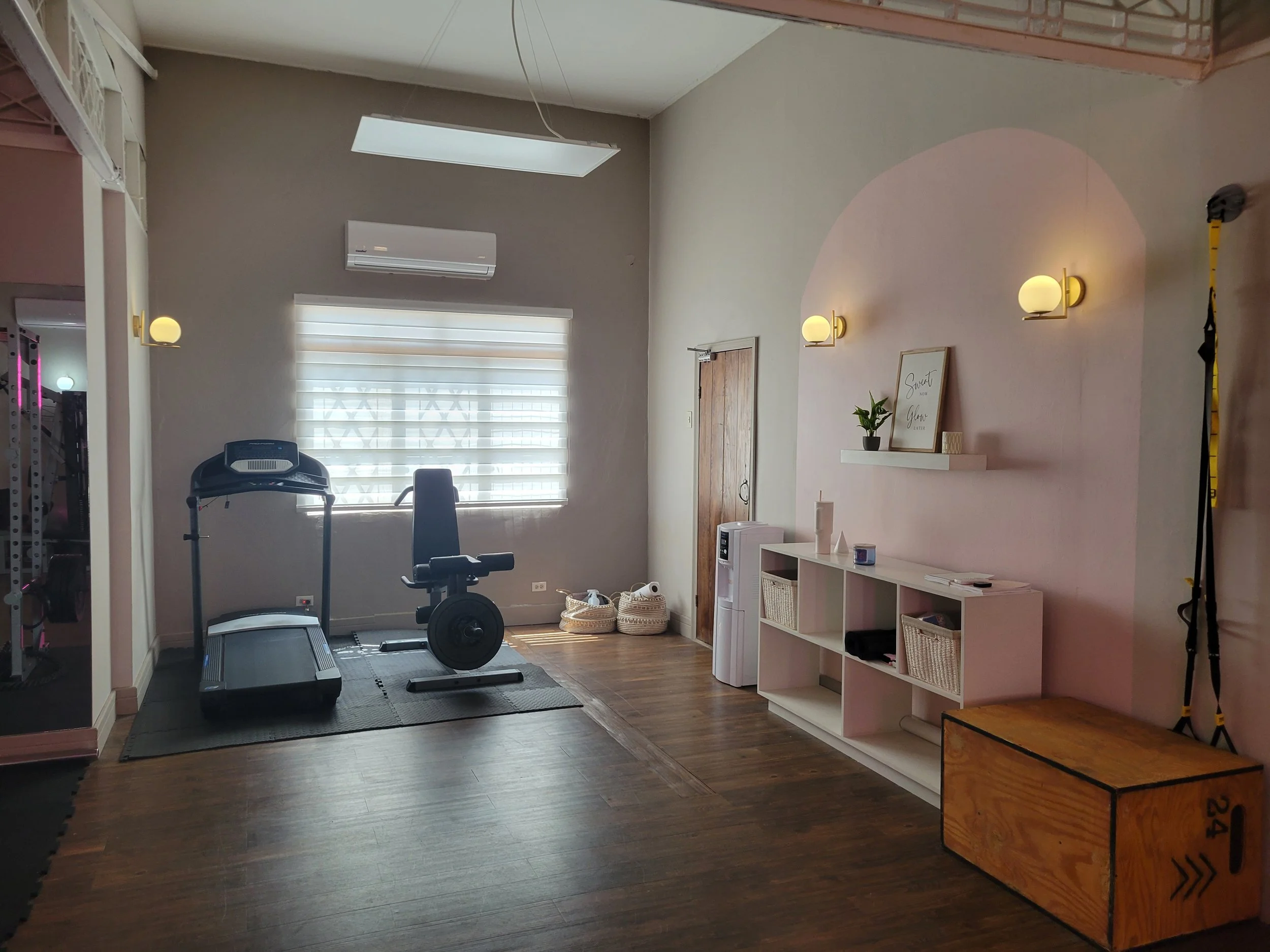 A home gym featuring a treadmill, a stationary bike, and a window with blinds, with a wooden box and a white storage unit with baskets and decor items on a pink wall.
