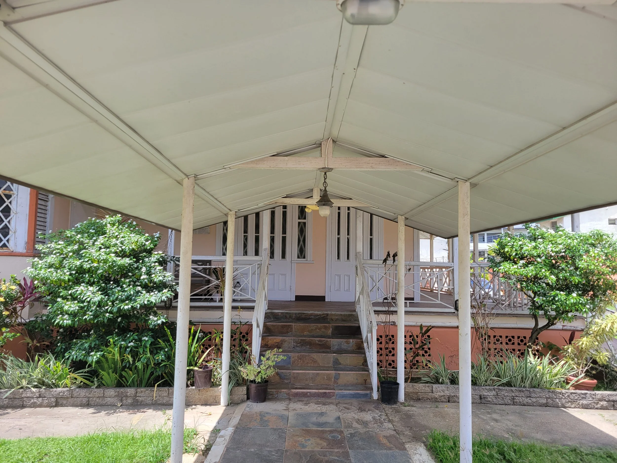Front porch of a house with white railing and stairs, surrounded by green plants and trees, with a covered roof and a ceiling fan.