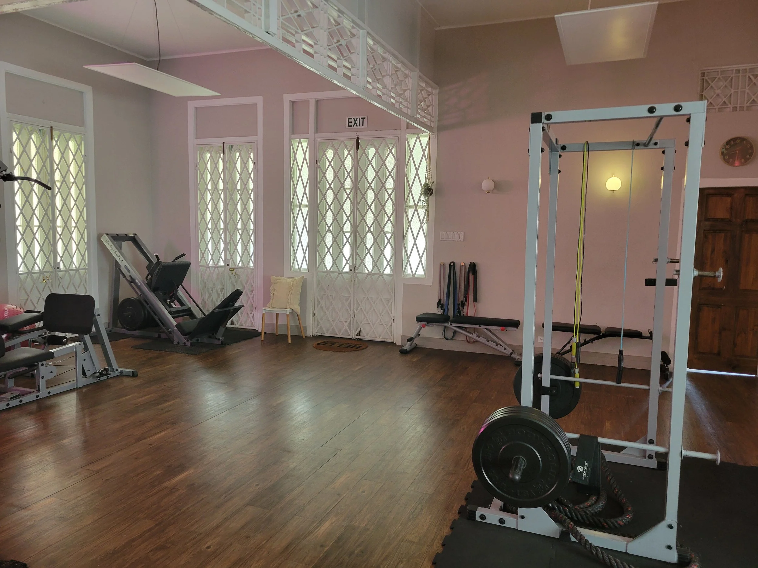 Empty gym room with workout equipment, including a rowing machine, benches, resistance bands, a squat rack with weights, and a wooden chair near windows with decorative screens.