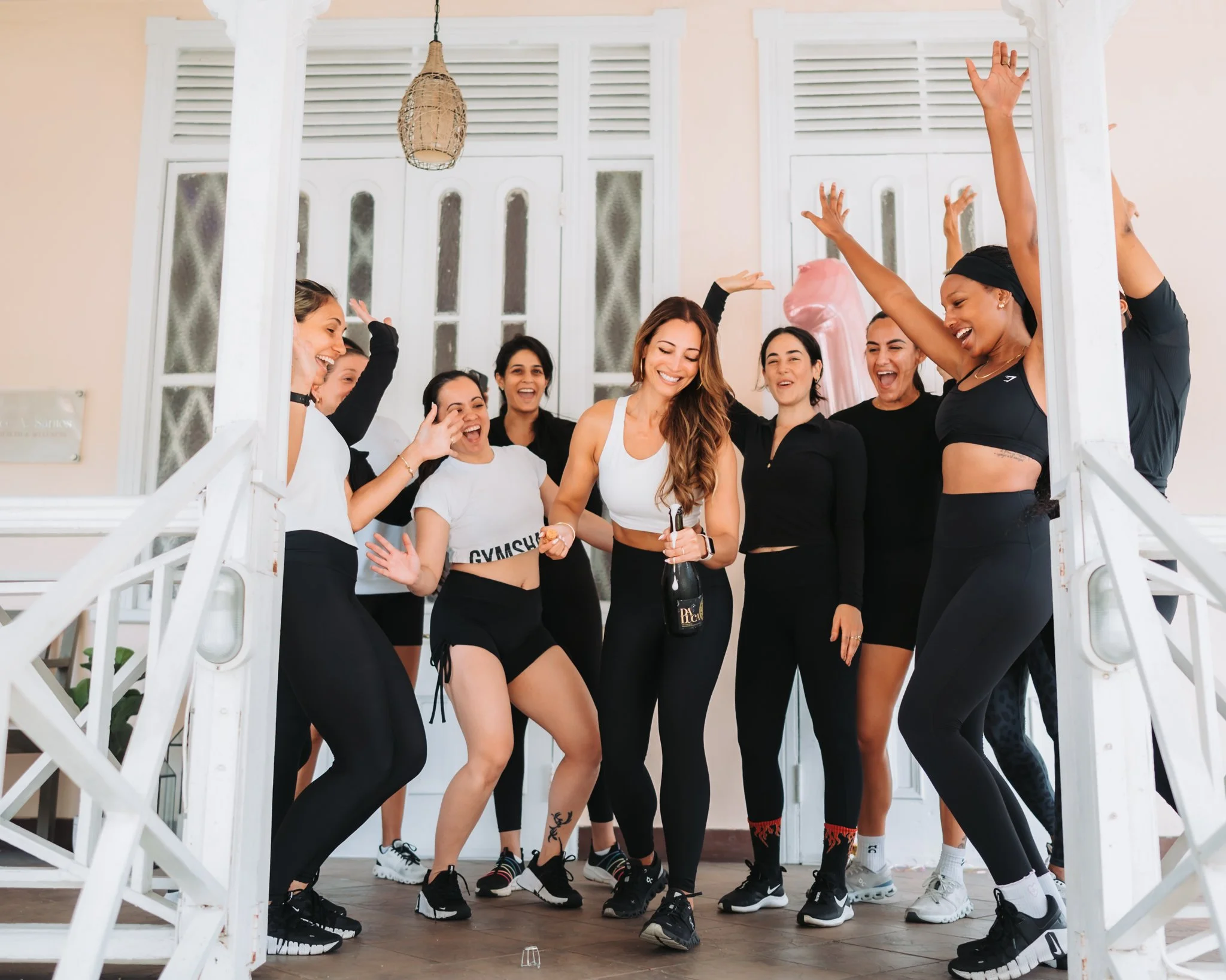 Group of women celebrating and dancing on a porch, with some holding drinks, wearing athletic and casual clothing, smiling and having fun.