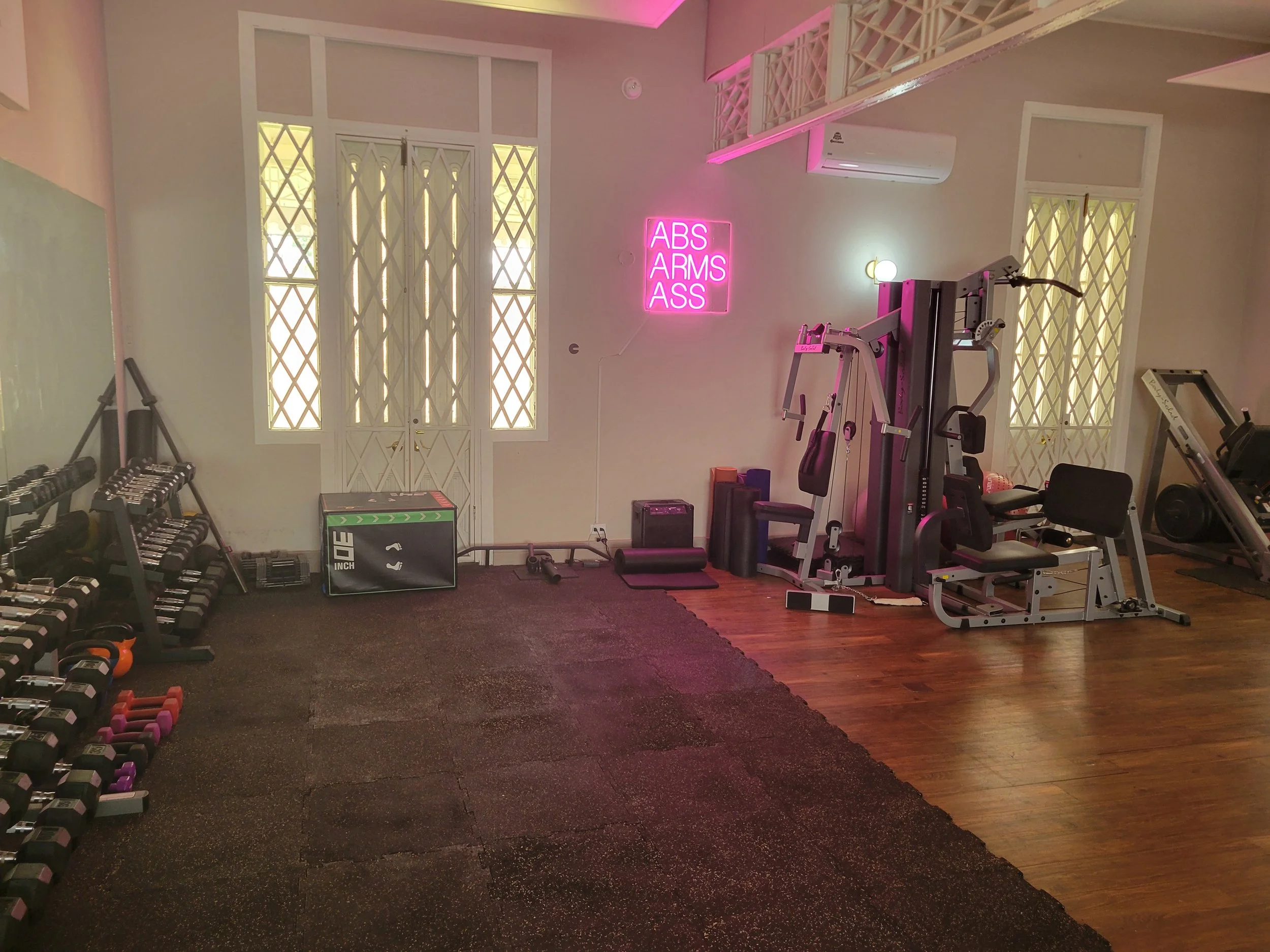 Gym room with dumbbells on racks, workout equipment, and a neon sign on the wall that reads "ABS ARMS ASS".