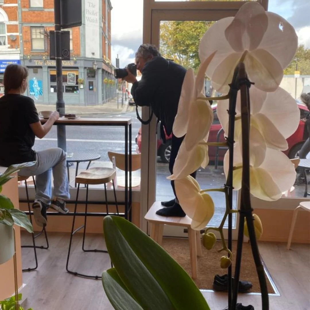 Photographer standing on a chair to photograph woman looking out the window drinking coffee