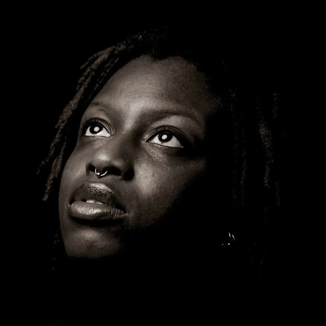 Black and white portrait of a person with a septum piercing, looking upwards against a dark background.