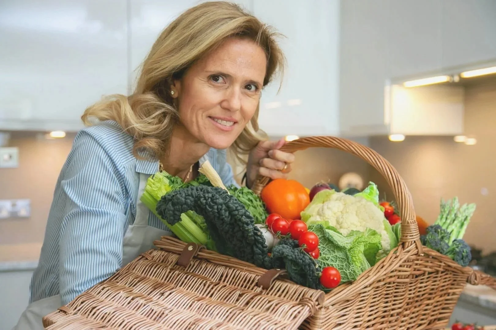Kensington Brand shoot ,Woman wsmiling and holding a  basket filled with fresh vegetables, in a kitchen.
