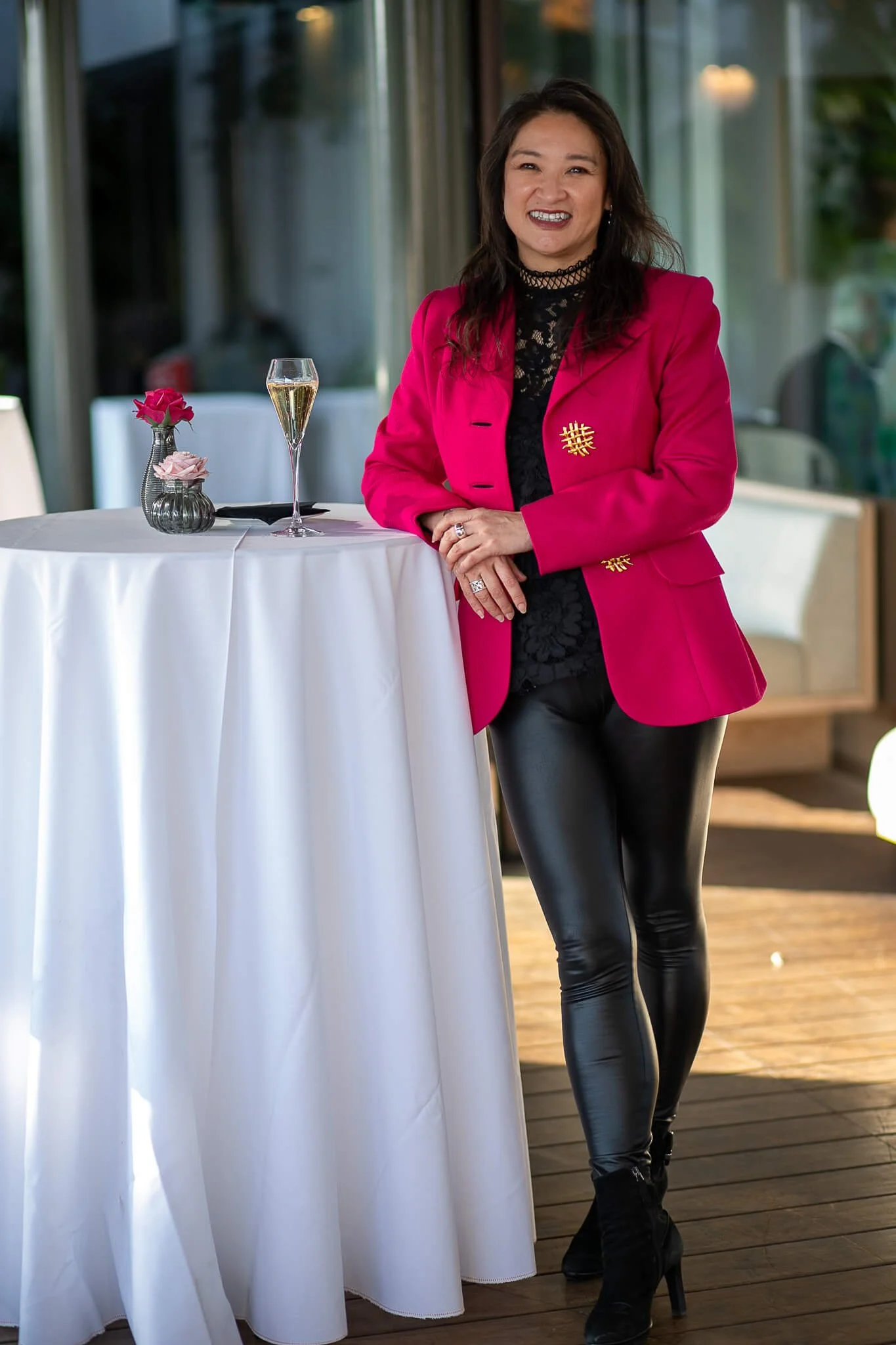 Full length shot of an asian woman in a pink jacket leaning against a table with  a glass of champagne and flowers on it