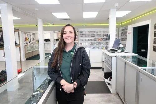 A young woman with long dark hair smiling inside a pawn shop, wearing a dark jacket and green shirt.