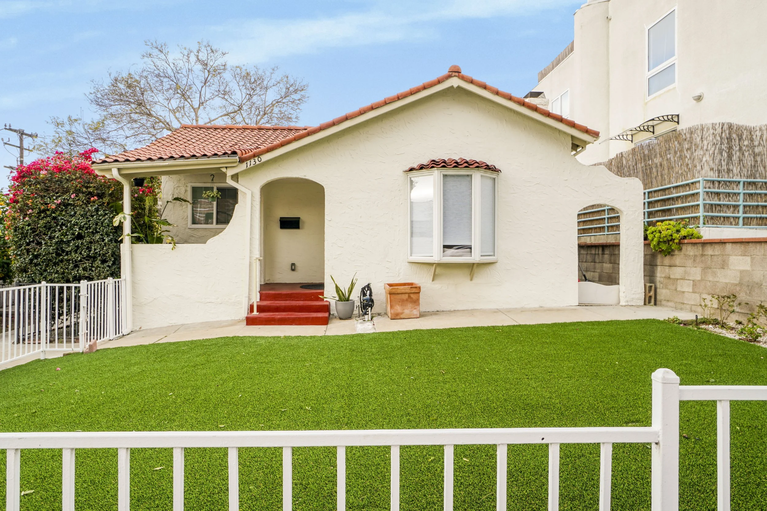 A small white stucco house with a red tile roof and a front lawn of bright green grass, enclosed by a white picket fence, with a sidewalk in front.